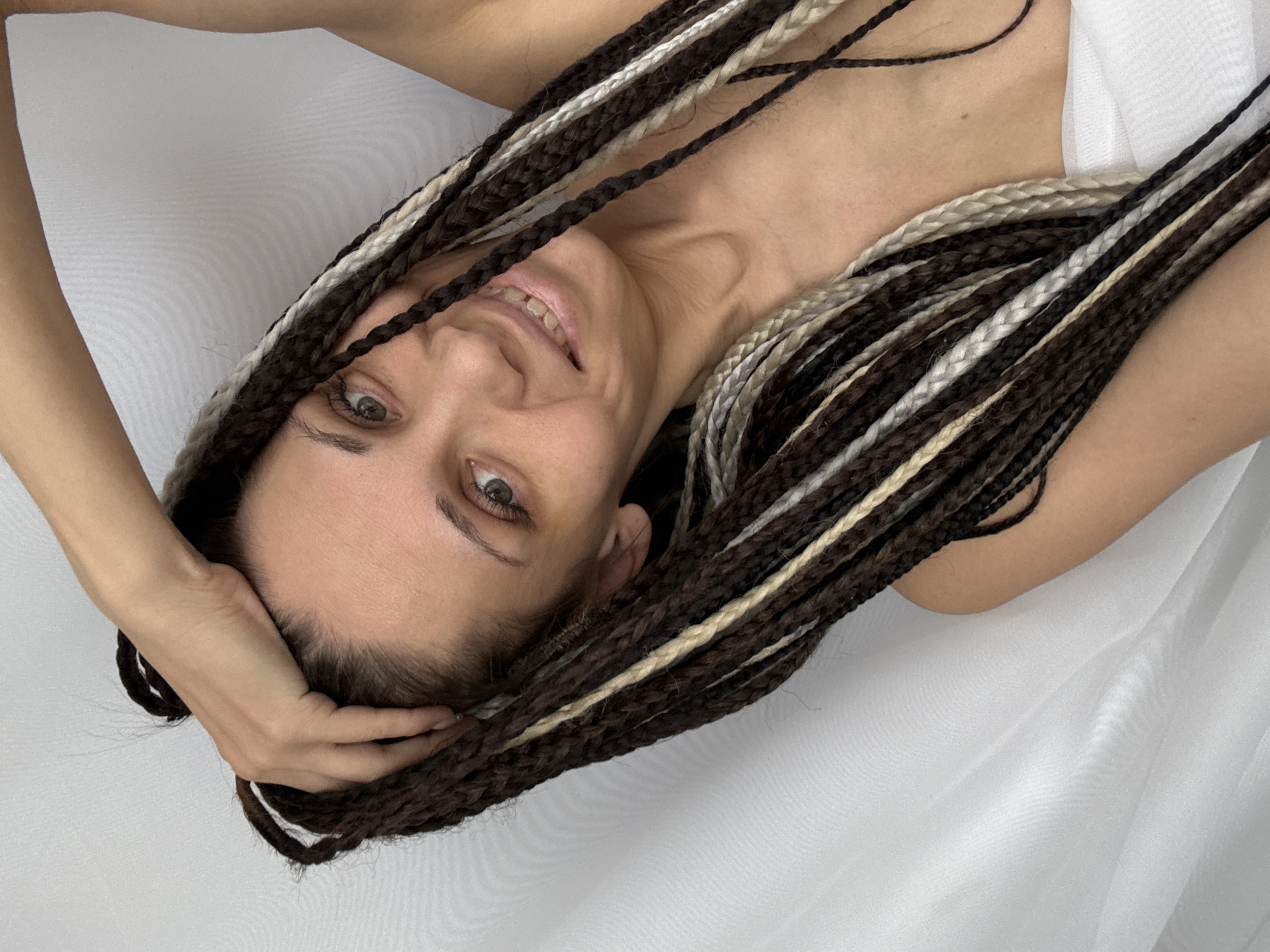 A woman with brown and blonde braided hair is lying on a white surface. Her left hand is placed on her head. She is smiling and looking at the camera. The background is a plain white surface. The location and city, Rusanovo, Russia, are not discernible from the image.