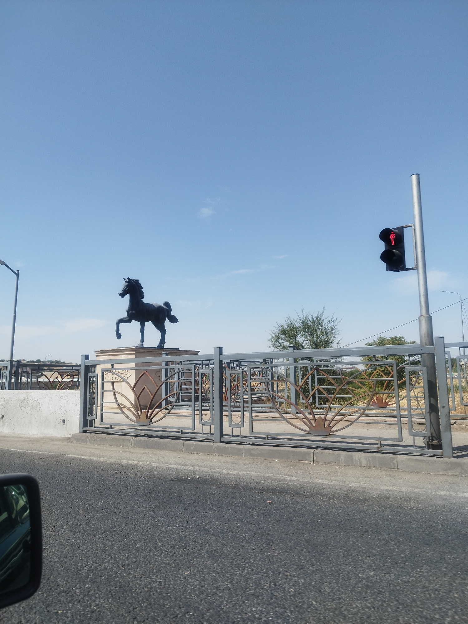 The image shows a statue of a horse on a pedestal, located in Shymkent, Kazakhstan. The statue is dark in color and appears to be made of metal. It is positioned behind a decorative metal fence with a floral motif. To the right of the statue, a traffic light is visible, displaying a red pedestrian signal. The background shows a clear blue sky, some trees, and a street in the foreground, suggesting an outdoor urban environment. The perspective suggests the photo was taken from a moving vehicle.