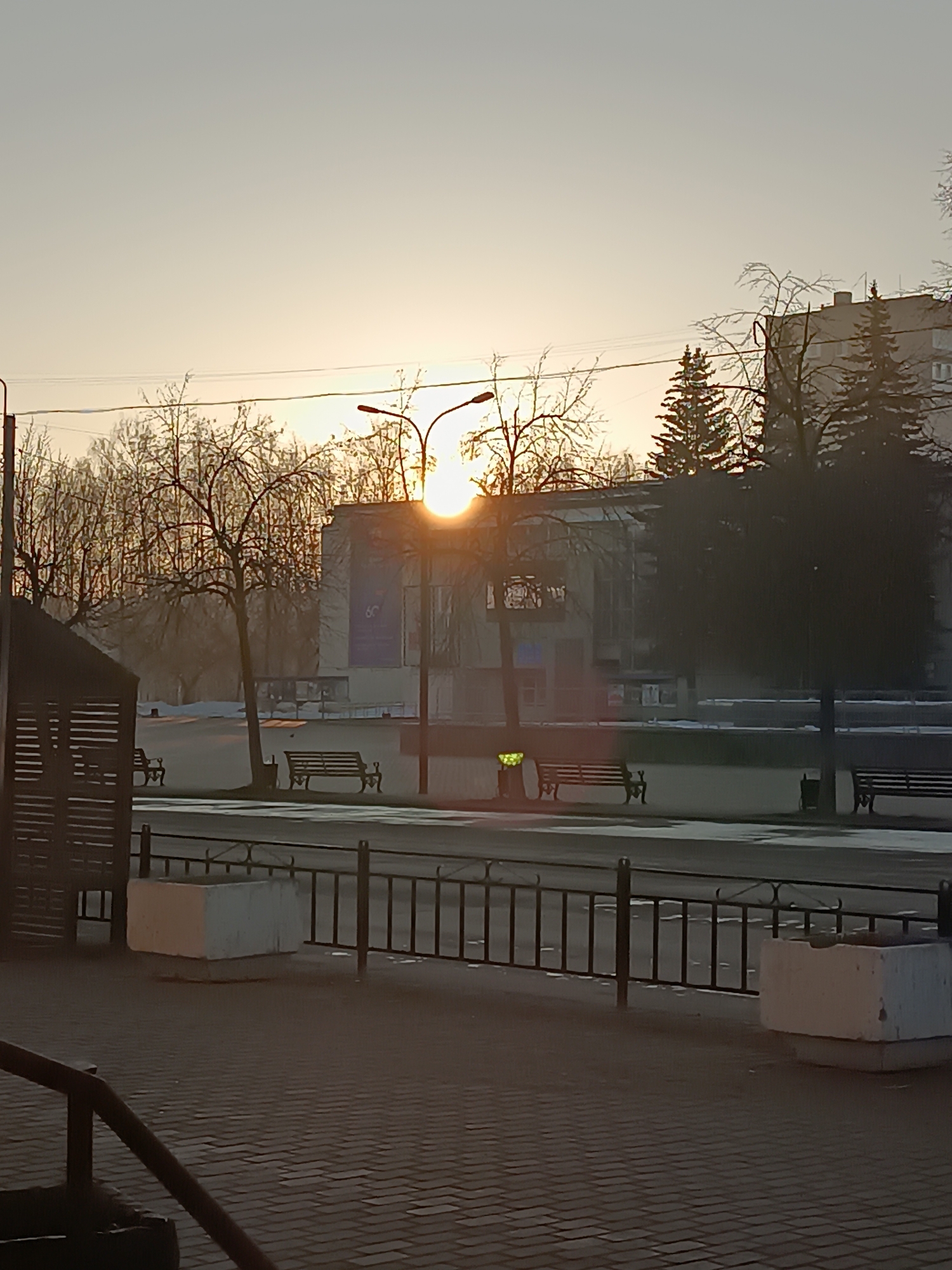 The image depicts a park scene in Chaikovsky, Russia, at sunrise or sunset. The sun is positioned behind a building, creating a silhouette effect. Several bare trees and evergreen trees are visible in the background. Benches are scattered throughout the park area. A black metal fence runs along the foreground, with white concrete planters positioned intermittently. The ground is covered in brick pavers. A partial structure, likely a bus stop, is visible on the left. The overall scene suggests a quiet and serene moment.