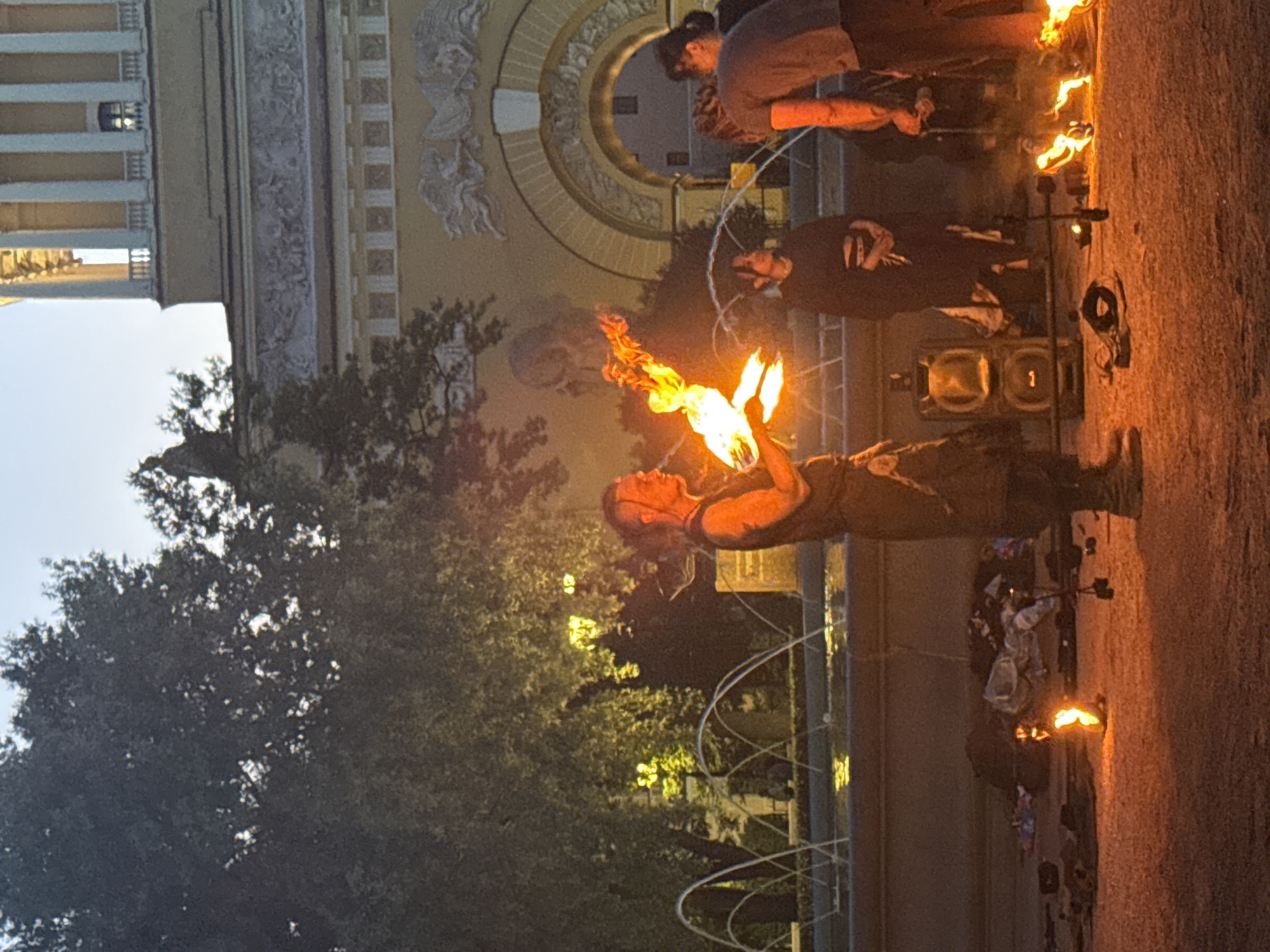 In Saint Petersburg, Russia, near the Field of Mars, a fire performance is taking place at dusk. The central figure, a man with his hair pulled back, is actively performing with fire, holding lit props that emit bright flames. He is dressed in dark, rugged clothing, possibly a vest and pants, with visible tattoos on his arm.
Several other individuals are observing the performance, some standing and others seated. One woman in the background is wearing a dark outfit and appears to be watching the fire artist. To the right, two other individuals are present; one is wearing a gray t-shirt and dark pants, and the other is partially obscured but also appears to be observing.
The environment features a fountain with multiple arching water jets in the mid-ground. Behind the fountain and the performers, a large, ornate building with classical architecture is visible, characterized by columns and detailed facades. Trees with dense foliage are scattered throughout the scene, some illuminated by artificial lights, creating a warm glow. The ground is covered in sand or gravel, with several objects, including what appears to be a speaker and various unidentifiable items, scattered around the performance area. Small fires are also burning on the ground in front of the performers. The sky is a soft blue, indicative of twilight.