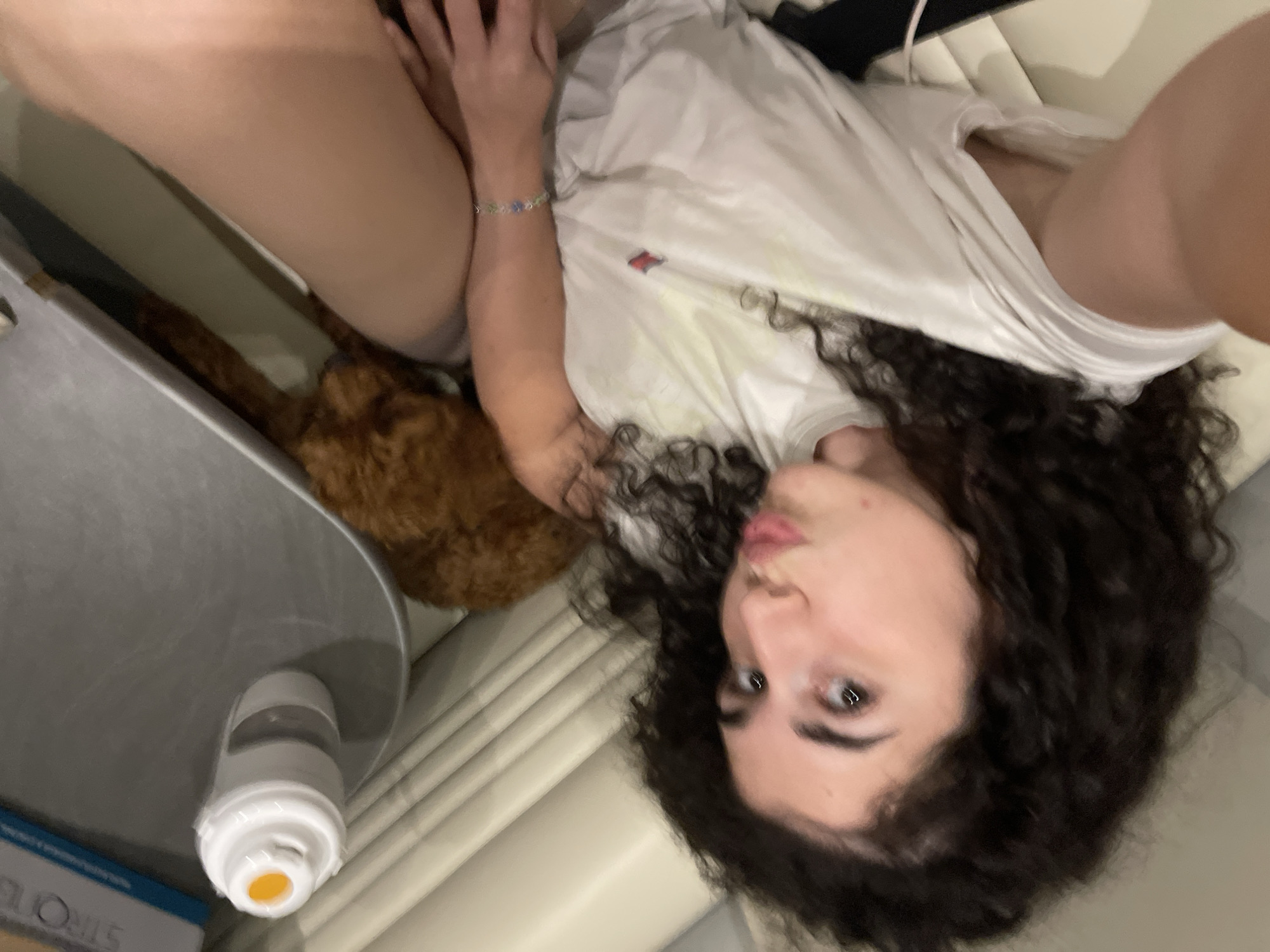 This image, taken in Koprinka, Bulgaria, depicts an indoor scene featuring a young person and a small dog. The person, likely female, has dark, curly hair and fair skin, and is lying down, facing the camera with a puckered lip expression. She is wearing a white t-shirt with a small red logo on the left chest and a colorful beaded bracelet with blue and green elements on her left wrist. A bare right leg (thigh) is partially visible. To her right, a small, fluffy, reddish-brown dog, possibly a poodle, is resting, mostly obscured by her leg and arm. The environment includes a white, ribbed surface to the left, and a grey, flat surface holding a white plastic bottle with an orange cap and a light blue box with the partial text "STRO" visible. The overall setting suggests a relaxed indoor space, possibly a living room or bedroom.