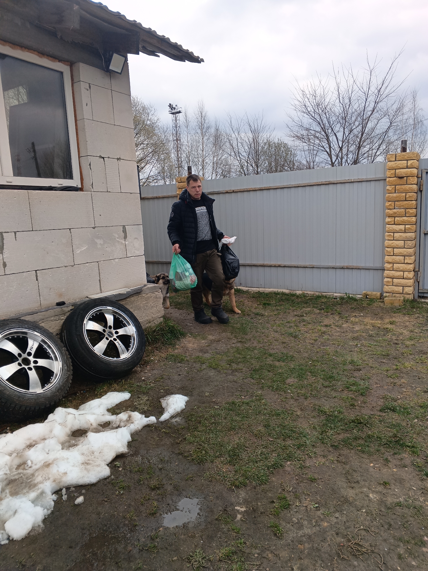 The image depicts a man standing in a yard in what appears to be Andreyevo, Russia. He is wearing a dark jacket, patterned sweater, and dark pants with boots. He is holding a green plastic bag in his right hand and a dark bag in his left. Beside him is a dog, possibly a German Shepherd, partially obscured by his legs and the bags he carries.
To the left of the man are two tires with silver rims leaning against the foundation of an unfinished building constructed of light-colored blocks. A window is visible in the building. Remnants of snow are on the ground near the tires.
The yard is grassy with areas of bare earth and puddles, suggesting a recent thaw or wet conditions. Behind the man is a grey fence with a brick pillar on the right side. In the distance, bare trees and a utility pole are visible against an overcast sky.