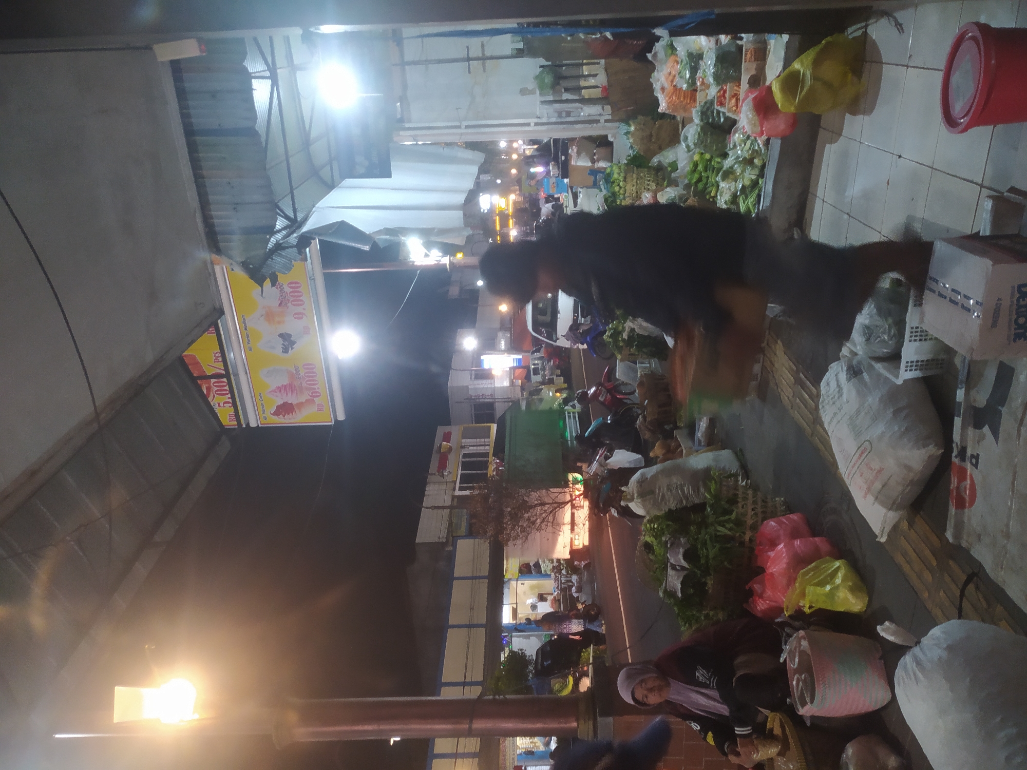 The image depicts a street scene at night, likely a market in Kupang, Indonesia. A prominent street lamp on the left casts a warm glow. In the foreground, a person in dark clothing is partially visible, facing away from the camera, with a box in their hands. To their right, a large red bucket is visible. A woman wearing a hijab sits on the left, near a woven mat with some items on it. Various bags, including pink and yellow ones, are scattered around. In the middle ground, a variety of produce, including leafy greens, cucumbers, and carrots, are displayed in baskets and on the ground. A white van and several motorcycles are parked on the street. In the background, buildings with lit windows suggest shops or residences. A large green truck is also present. A banner with images of ice cream and prices in Indonesian Rupiah is visible on a stall. The overall atmosphere is that of a bustling night market, with goods for sale and people engaged in commerce.
