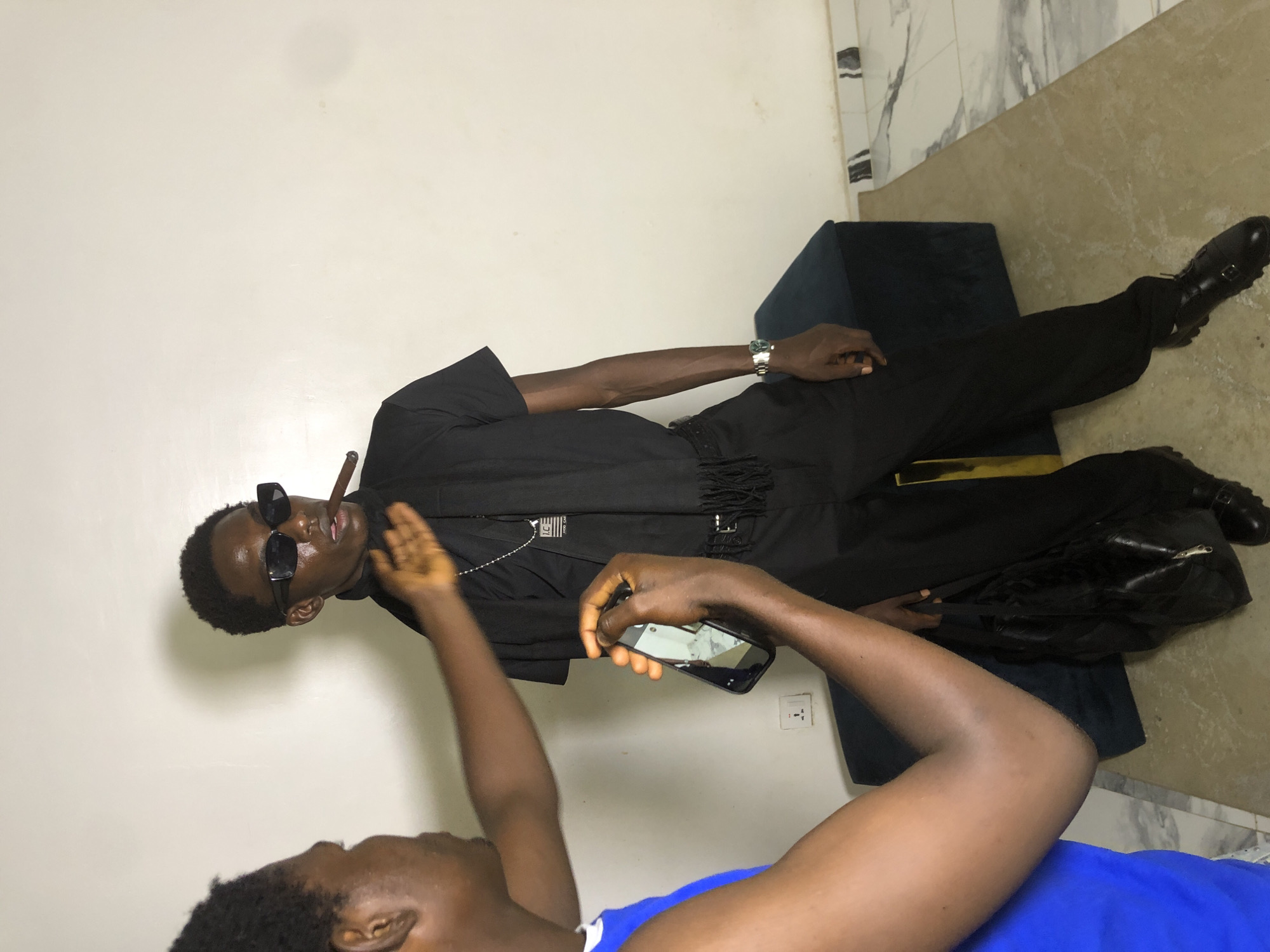 Two individuals are present in an indoor setting, possibly a room in Ado, Nigeria. The primary subject, a young man, stands facing towards the right of the frame. He is dressed in a black t-shirt, black pants, and black shoes with double buckles. He wears black rectangular sunglasses and has a cigar in his mouth. Around his neck, he has a black scarf and a silver chain with a pendant. A silver watch is visible on his left wrist. He is standing next to a dark blue bench or ottoman.
To the left of the primary subject, another young man is partially visible, facing the primary subject. He is wearing a blue sleeveless shirt. His right arm is extended towards the primary subject, with his hand open, as if interacting or gesturing. He is holding a smartphone in his left hand, which appears to be recording or taking a picture. A black bag is hanging from his right arm.
The background consists of a plain, off-white wall and a tiled floor with a marble pattern. A white electrical outlet is visible on the wall. The lighting appears to be artificial, casting soft shadows. The overall impression is a candid moment, possibly a photoshoot or casual interaction.
