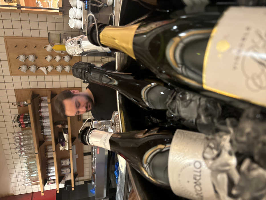 A man in a black shirt stands behind a metal bar, near shelves stocked with bottles and glasses. In front of him, champagne bottles are submerged in ice inside a container. A coffee machine is on the counter next to the champagne. Small white cups hang on a wooden rack on the wall behind the bar. The image is taken at an oblique angle, emphasizing the bottles in ice.