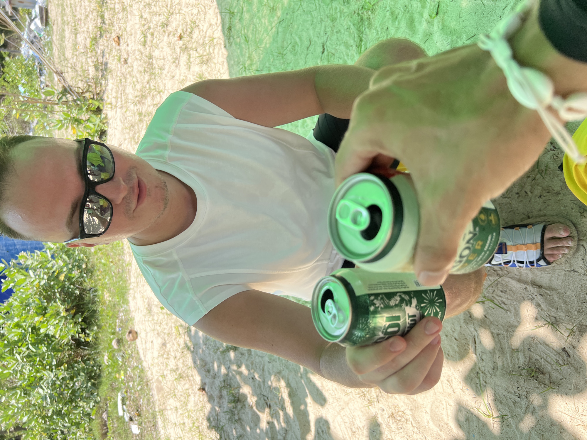 The image captures an outdoor interaction between two individuals on a sandy terrain in Dai Lanh, Vietnam. In the mid-ground, a light-skinned male with short hair, wearing reflective black sunglasses, a white sleeveless top, black shorts, and white, blue, and orange sandals, is crouched. He extends a green aluminum can, identified as "Bia Saigon," with his left hand towards the camera.
In the immediate foreground, a second person's right hand and forearm are visible on the right side of the frame, holding another identical green "Bia Saigon" can, which is positioned as if clinking with the can held by the first individual. A white, shell-like bracelet adorns the wrist of the foreground person. A yellow object is partially visible on the ground at the bottom right.
The environment consists of light-colored sand with scattered patches of green grass and small, dried debris. In the background, dense green foliage is prominent, with a blue tarp or fabric structure visible behind the greenery on the upper left. The lighting suggests a bright, sunny day with shadows indicating overhead cover.