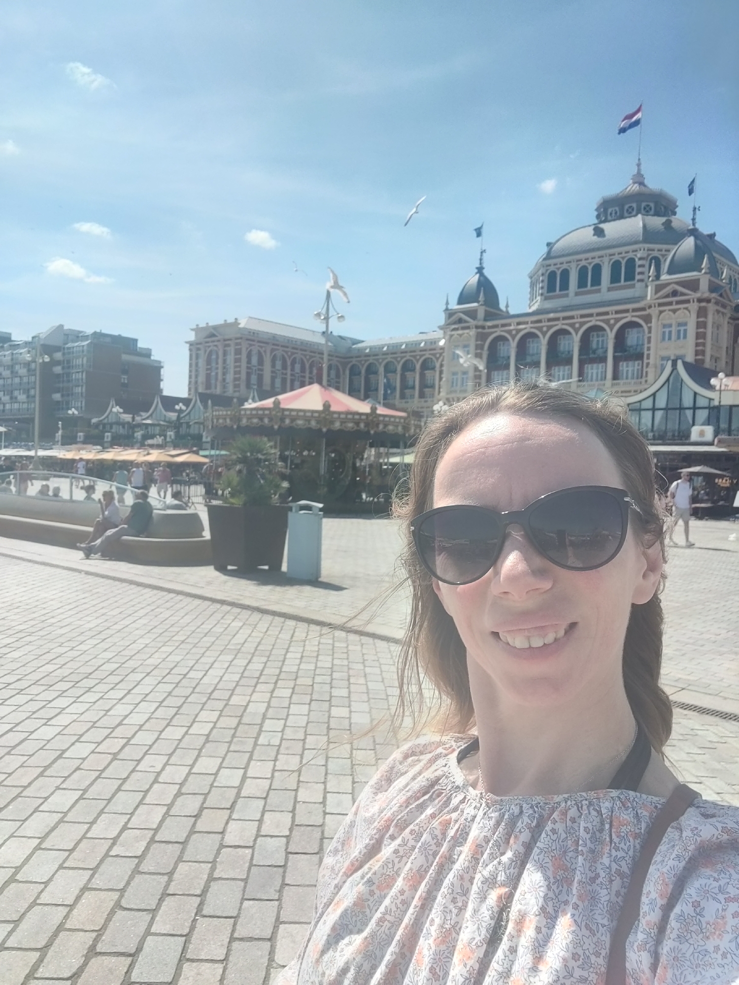 A medium shot selfie captures a woman with shoulder-length brown hair and sunglasses, smiling at the camera. She is wearing a floral-patterned blouse. Behind her, a grand building with a domed roof and Dutch flags is visible. Several seagulls are flying in the sky. The scene appears to be a waterfront promenade in The Hague, Netherlands, with paved ground and other buildings in the background. In the middle ground, a carousel and people sitting on a bench add to the atmosphere of a public space.