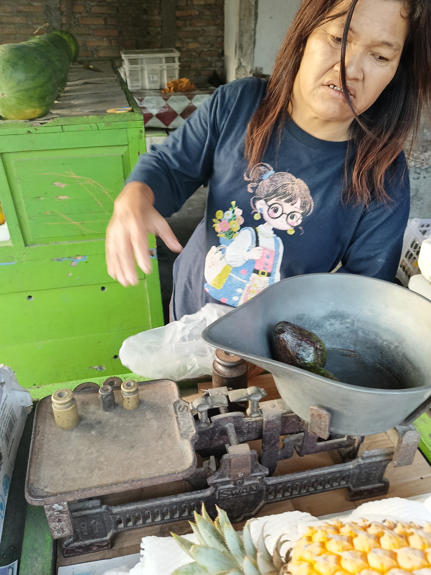 In Borongan, Indonesia, a woman with dark hair and glasses, wearing a dark blue t-shirt with a cartoon image, is weighing an avocado on an old, rusted mechanical scale. The scale has a flat, square platform on the left with brass weights and a wider, grey basin on the right, where the avocado rests. The woman's left hand is blurred as it moves, suggesting she is in the process of the weighing. Behind the scale, a plastic bag is visible. The scene appears to be an outdoor market stall, with a bright green wooden counter or structure to the left and a brick wall in the background. To the left of the counter, a large, green, oblong object, possibly a melon or squash, is visible. In the foreground, parts of a pineapple and another fruit are visible, suggesting a fruit stall. The lighting indicates it is daytime.