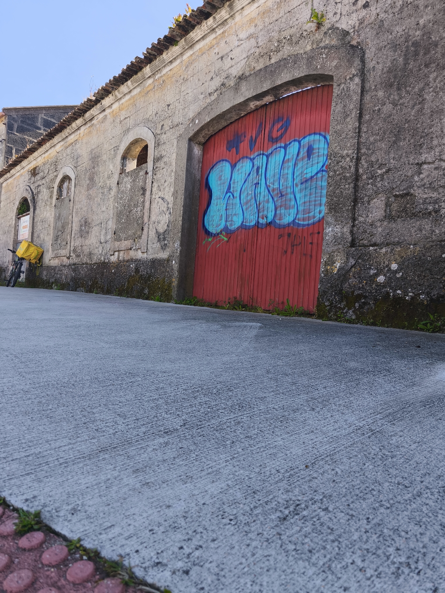 Captured in Cangas de Morrazo, Spain, the image presents a street scene. A gray stone building stretches along the left side, featuring a red-painted wooden door with blue graffiti and arched windows. A bicycle equipped with a yellow delivery bag is parked near the building. The foreground is dominated by a sloping gray concrete sidewalk with a red brick border visible at the bottom left. The sky above is light blue. The building's stonework shows signs of age and wear, with moss and discoloration present.