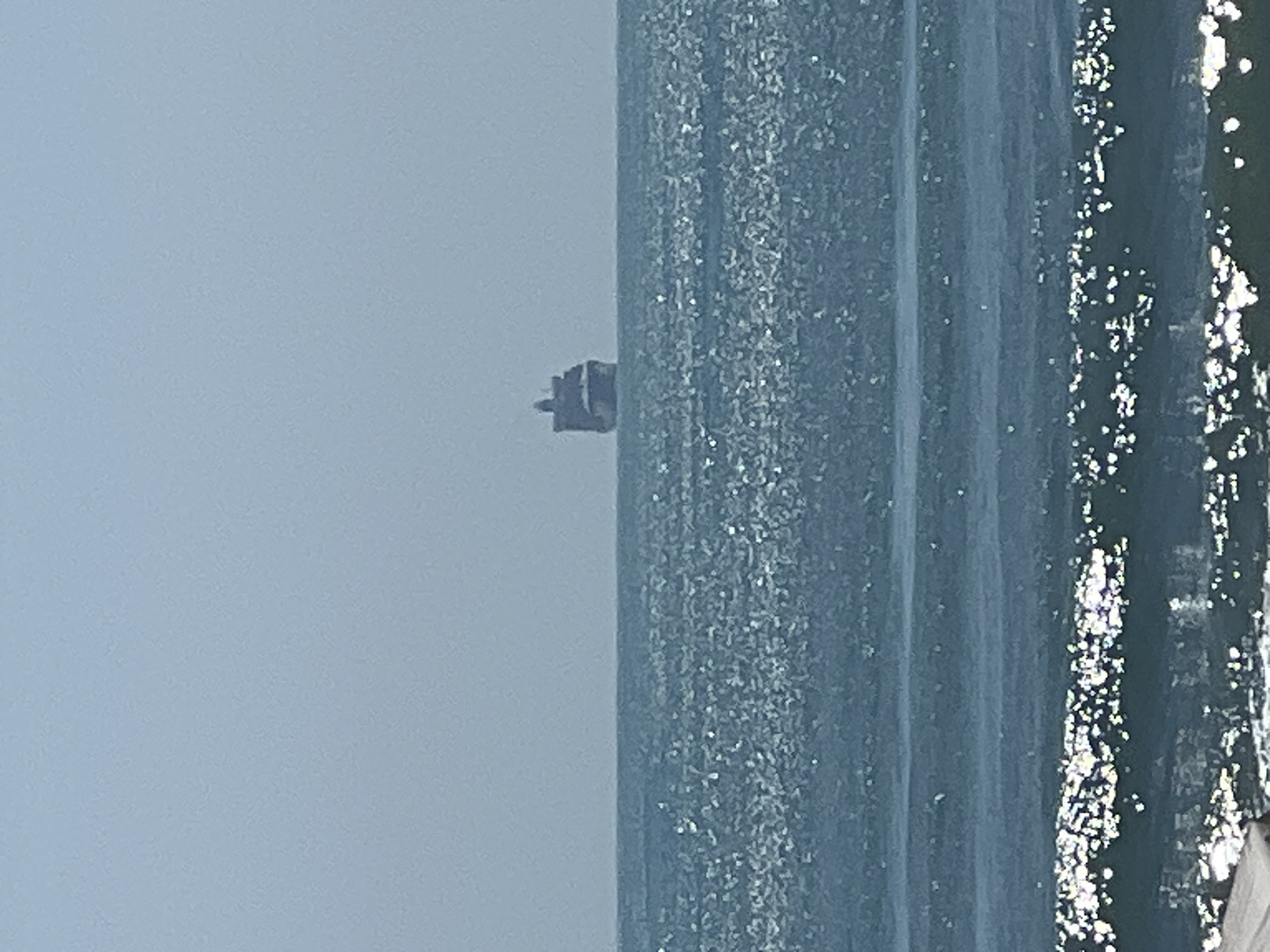 The image displays an expansive seascape under a pale, light blue-grey sky. The horizon line clearly separates the sky from the vast body of water, which dominates the lower and right portions of the frame (when correctly oriented).
The sky is largely featureless, suggesting an overcast or hazy daytime condition. The surface of the water exhibits varying textures and colors; it is predominantly blue-grey with numerous bright, sparkling white reflections scattered across its surface, especially towards the right. These glints indicate sunlight reflecting off ripples or small waves, confirming it is daytime. Some areas of the water appear as smoother, darker blue vertical bands.
In the distance, positioned on the horizon against the pale sky, is a small, dark, and somewhat indistinct object. Its silhouette is blocky and appears to have multiple levels, resembling a distant ship or a small offshore structure. The object is not clearly defined due to its distance from the viewer.
There are no people visible, and no specific activities are taking place. The scene conveys a sense of openness and tranquility. The visual cues suggest a coastal view, consistent with a location such as Varna, Bulgaria, overlooking the Black Sea. No visible text is present.