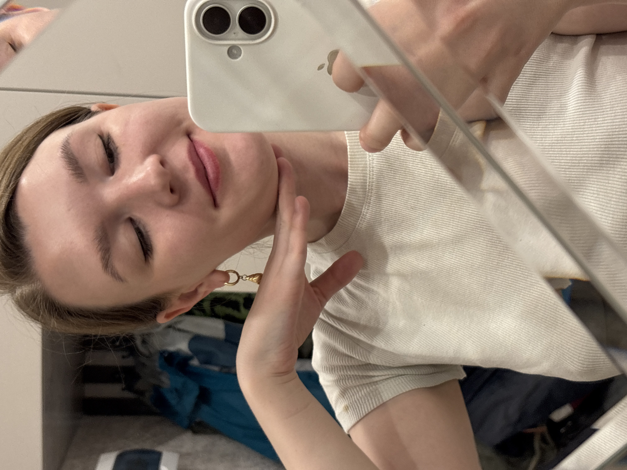 A woman is taking a selfie in a mirror. She is holding a white smartphone, likely an iPhone, close to her face. She is wearing a white ribbed t-shirt. Her hair is blonde, and she has a gold hoop earring in her left ear. Her right hand is near her neck. The background is blurry and shows some clothes and furniture. The location is not discernible.