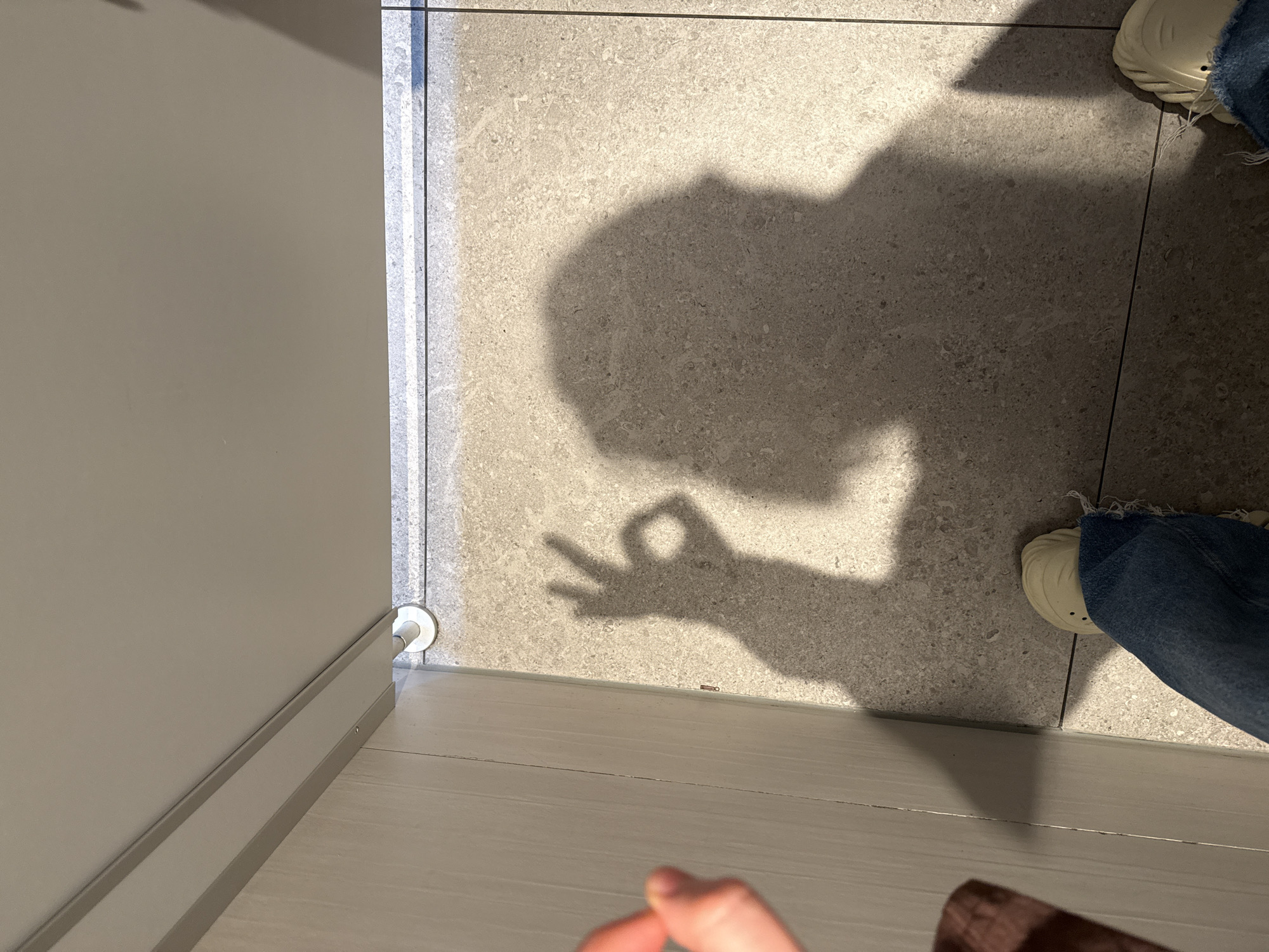 The image displays an indoor scene, likely in Waldshut, Germany, dominated by the shadow of a person cast upon a light-grey tiled floor. The shadow, occupying the upper right and central parts of the frame, distinctly shows the profile of a head and upper body, with one hand making an "OK" gesture.
To the right, the lower part of a person's leg clad in blue denim jeans and a light-colored, possibly beige, textured shoe is visible, partially obscured by the shadow. Another foot and leg are partially seen in the bottom right corner. The floor is covered with speckled, light-grey tiles with visible grout lines, suggesting a modern interior.
On the left side of the image, a plain light-grey wall stands vertically, meeting the floor with a matching baseboard. A small, white, circular object, possibly a floor-mounted doorstop or buffer, is visible where the wall meets the floor in the bottom left. In the immediate foreground, at the very bottom, out-of-focus fingers belonging to a hand are visible, suggesting the perspective of the photographer. A tiny metallic clip or staple lies on the lighter floor section near the baseboard.
The bright lighting creates sharp, well-defined shadows, indicating a strong light source, possibly natural light from a window or powerful artificial illumination. There is no visible text in the image. The scene captures a casual moment in what appears to be a contemporary building interior.