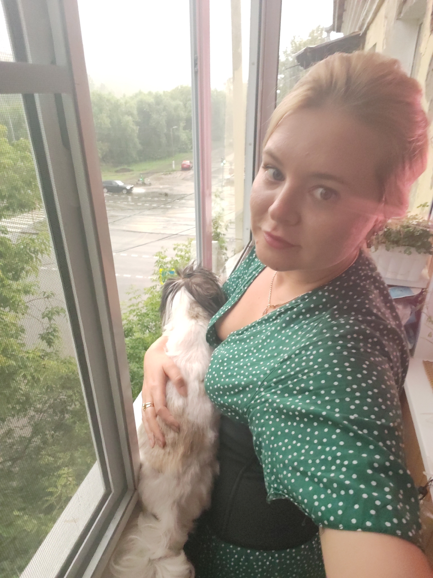 A blonde woman with pink-tinted hair is holding a fluffy, light-colored dog with dark markings in her arms. She is wearing a green dress with white polka dots and a gold necklace. The woman and dog are standing near a window, with a view of a street outside. The street appears to be wet, suggesting it might be raining or has recently rained. There are trees and buildings visible in the background. The window has a screen, indicating it might be a balcony or an open window. The overall impression is a casual, indoor portrait with an outdoor backdrop. The presence of a residential building and a street with a few cars suggests an urban environment within Izhevsk, Russia.