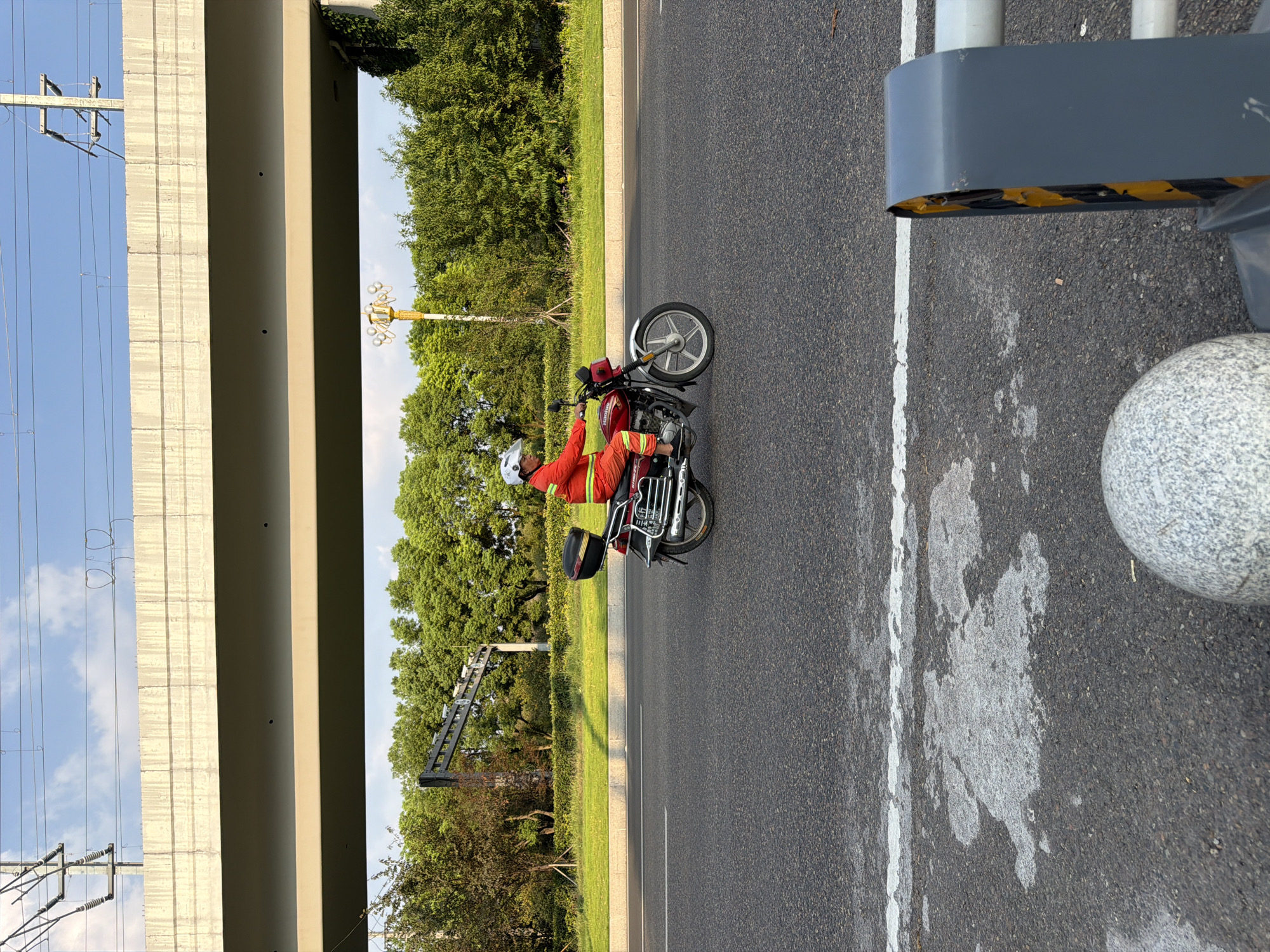 A person wearing a bright orange suit and white helmet rides a red motorcycle with a black storage box attached to the rear. The motorcycle is on an asphalt road with a white stripe marking the lane. In the background, there is a large overpass, a line of green trees, and a clear blue sky with some clouds. The scene appears to be in the city of Jiangjin, China, as indicated by the context.