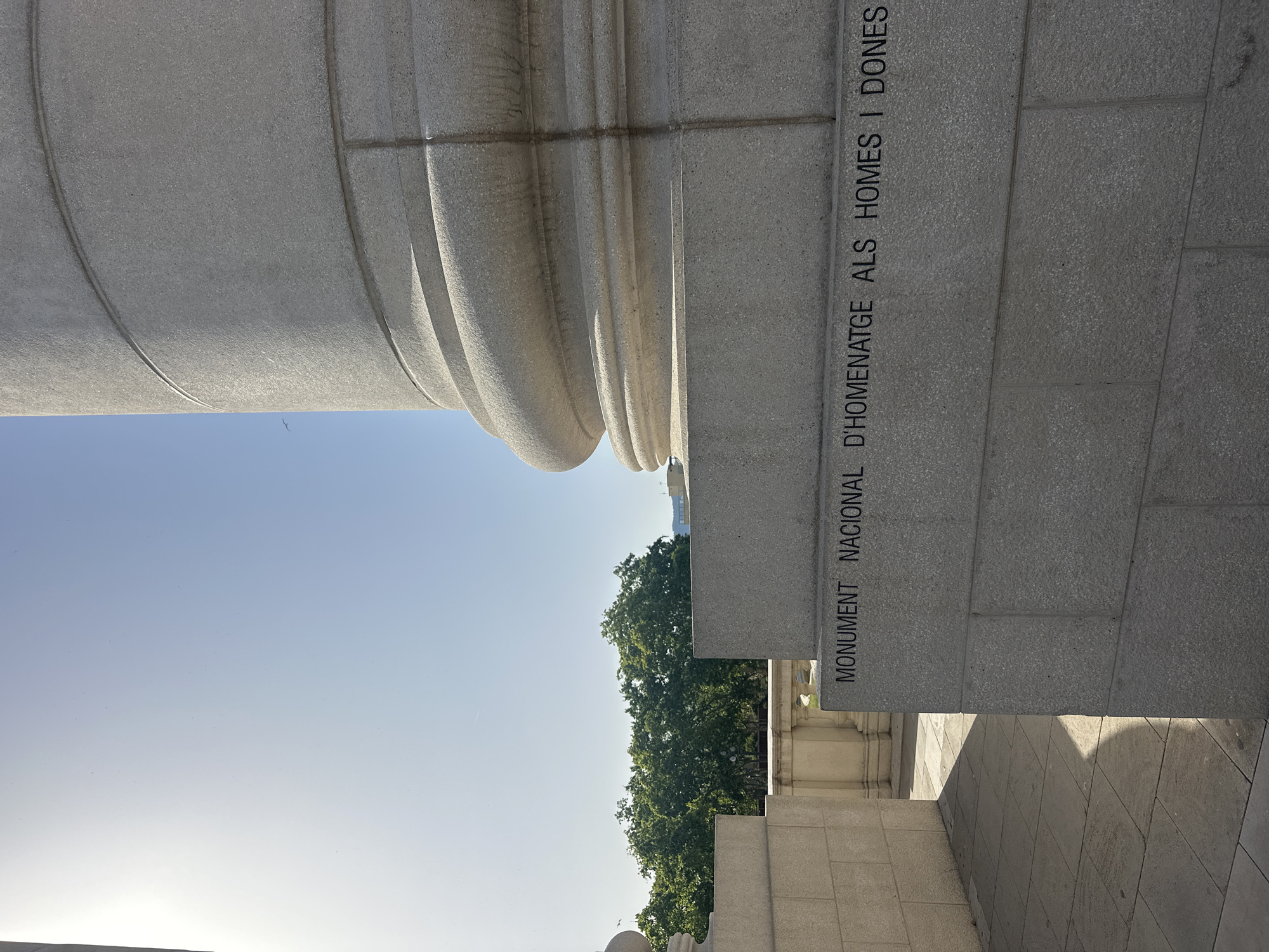 The image shows a close-up of a large stone column with inscription, set against a clear blue sky and some trees in the background. The inscription on the base of the column reads "MONUMENT NACIONAL D'HOMENATGE ALS HOMES I DONES". This monument is located in Barcelona, Spain. The scene is outdoors, with sunlight casting shadows on the stone paving in the foreground. There are no people visible in the image. The focus is on the architectural details of the monument and its inscription.