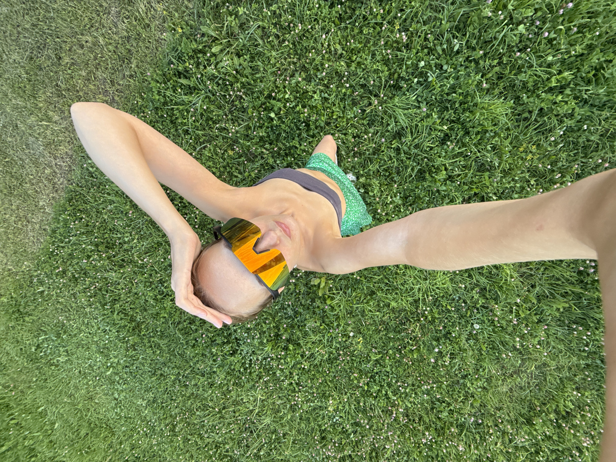 A person is lying on a grassy field in Podshivalovo, Russia, holding their head with one hand. They are wearing futuristic-style sunglasses with orange reflective lenses, a dark gray sports bra, and a green patterned skirt. The grass is green and speckled with small pink flowers. The image is taken from a high-angle perspective, looking down at the person and the surrounding field.