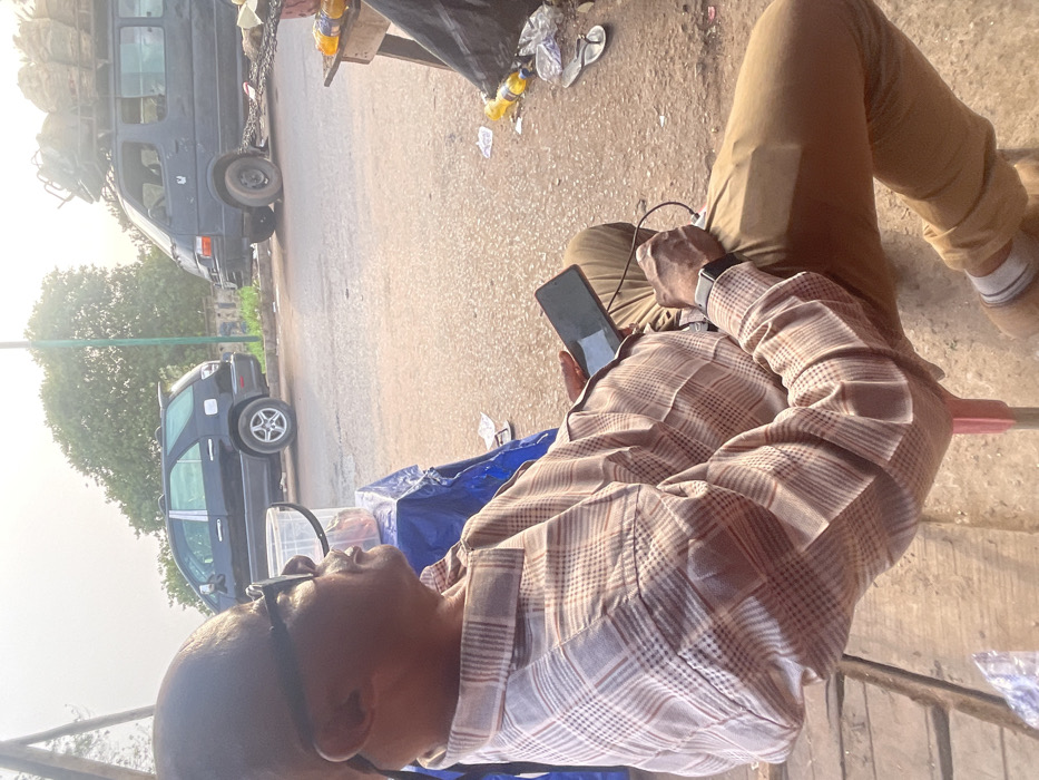 A man wearing glasses and a patterned shirt sits casually with his legs outstretched. He's holding a smartphone connected to a charger. Behind him is a street scene with several vehicles, including a van heavily loaded on its roof, parked along the roadside. Debris is scattered nearby, adding to the informal atmosphere.