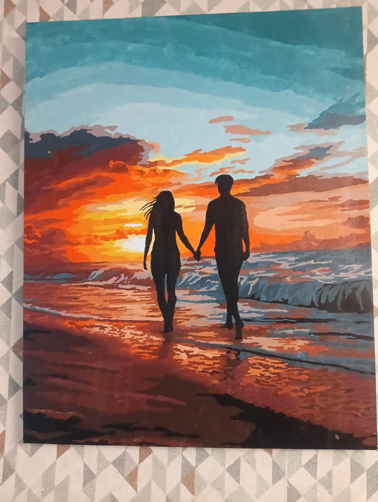 The image is a painting depicting a silhouetted couple walking hand-in-hand on a beach at sunset. The sky is a gradient of blue at the top transitioning to vibrant oranges, reds, and yellows near the horizon. The sun appears to be setting directly in front of the couple. Waves are breaking on the shore in the background. The sand reflects the colors of the sunset.
There is no evidence to suggest this painting is located in Vsevolozhsk, Russia. The scene depicts a beach, and Vsevolozhsk is inland.