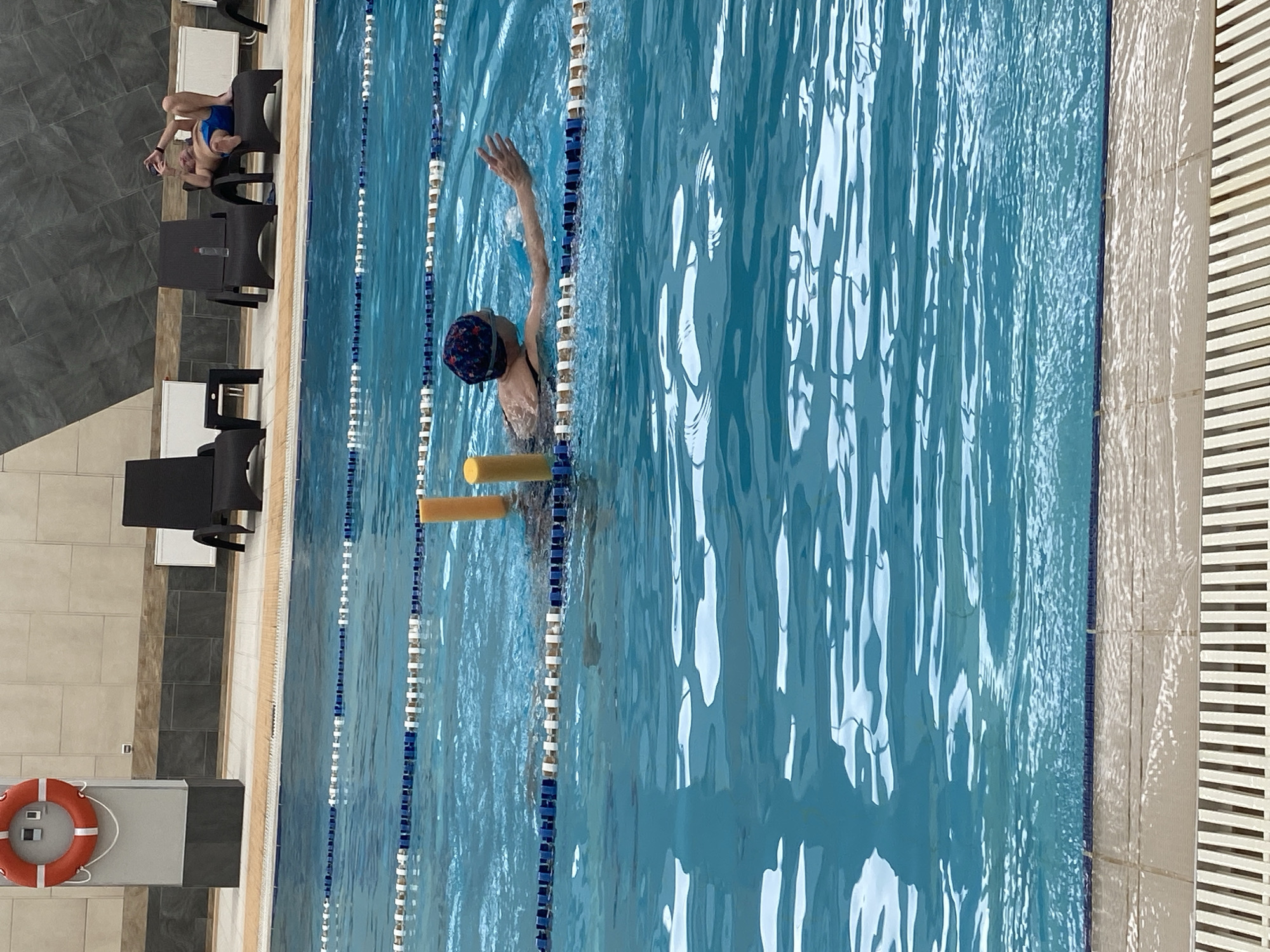 An overhead shot shows an indoor swimming pool, likely in Yekaterinburg, Russia, given the context. A woman in a swim cap is in the pool, using a foam flotation device. The pool is divided into lanes by blue and white lane markers. Seating is visible along the poolside. Another person is sitting and watching. A life preserver is mounted on a wall near the pool. The water is blue with light reflecting off its surface. The pool's edge is bordered by a tiled walkway and a white grated design.