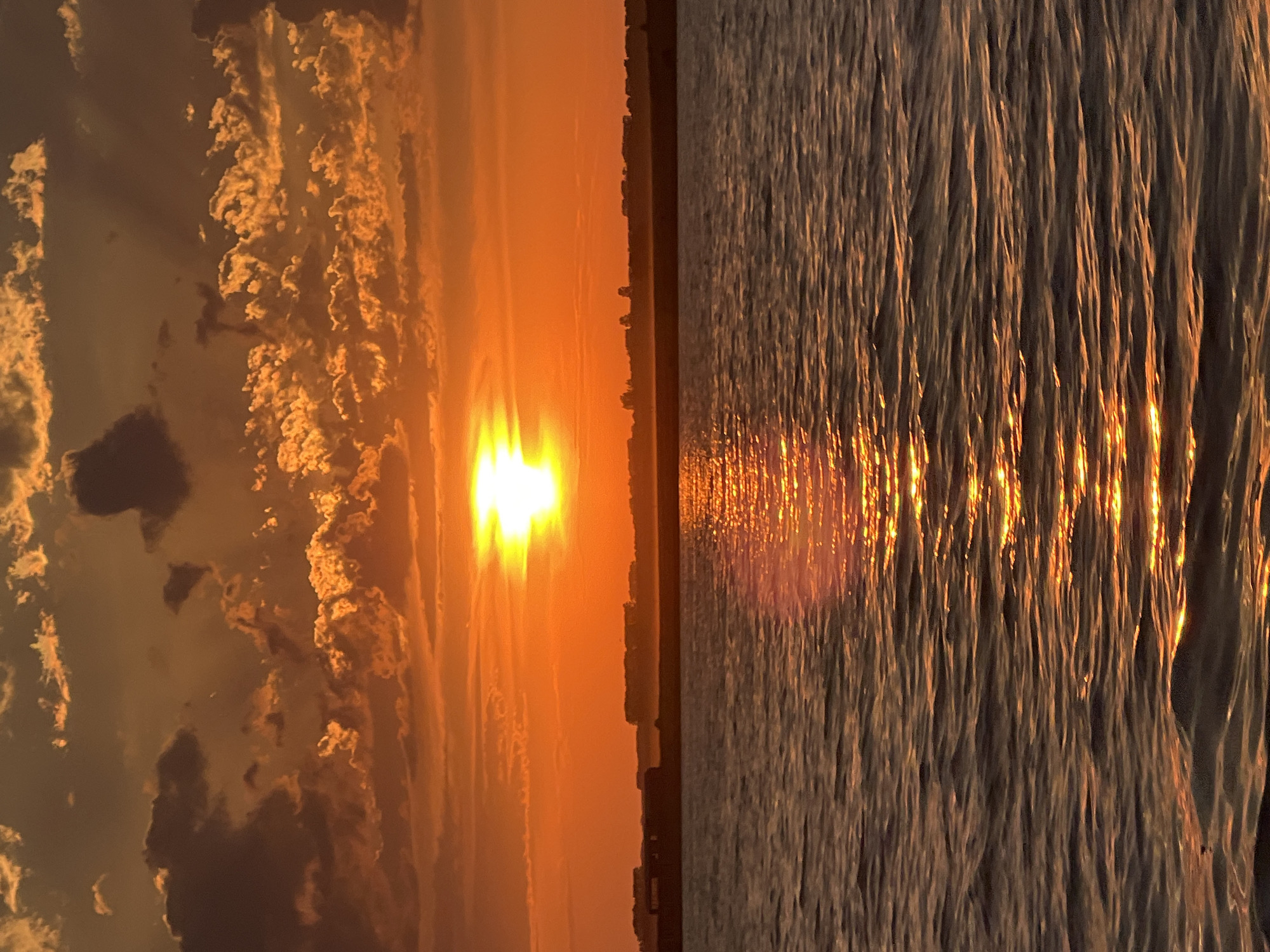 A vibrant sunset over a body of water is depicted. The sun, a bright white disc, is positioned slightly left of center in the upper portion of the frame, casting a warm orange glow across the sky. Scattered clouds, tinged with orange and brown, fill the sky, with some appearing darker and more defined. A thin, dark horizontal line, representing land or a distant shore, stretches across the middle ground. The water in the foreground is rippled, reflecting the warm hues of the sunset in shimmering streaks of orange and gold. The overall scene conveys a sense of tranquility and natural beauty. The location is identified as Novopichugovo, Russia.
