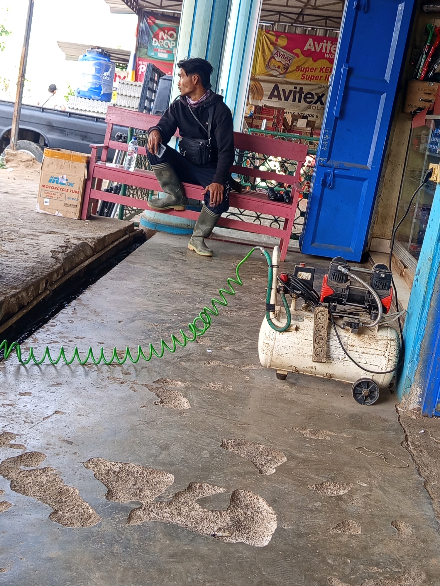 A medium shot captures a man sitting on a red bench outside a shop in Karang Sari, Indonesia. The man, wearing a black hoodie and grey rubber boots, looks to his right, his posture relaxed. Beside him on the bench rests a water bottle and a black bag. To his left, a cardboard box labeled "Motorcycle Tube" is visible.
In the foreground, a cream-colored air compressor with red accents is situated on the concrete ground. A coiled green hose extends from the compressor and disappears out of frame to the left. The ground itself is uneven, with several patched areas and a dark, wet channel running horizontally.
Behind the man and the bench, the shop's facade features blue and light-colored walls, with signage bearing the "Avitex" brand. A large blue door is prominent to the right. Further back, a large blue water tank is perched atop a structure. The overall environment suggests a repair shop or a roadside stall. The lighting is bright, indicating daylight.