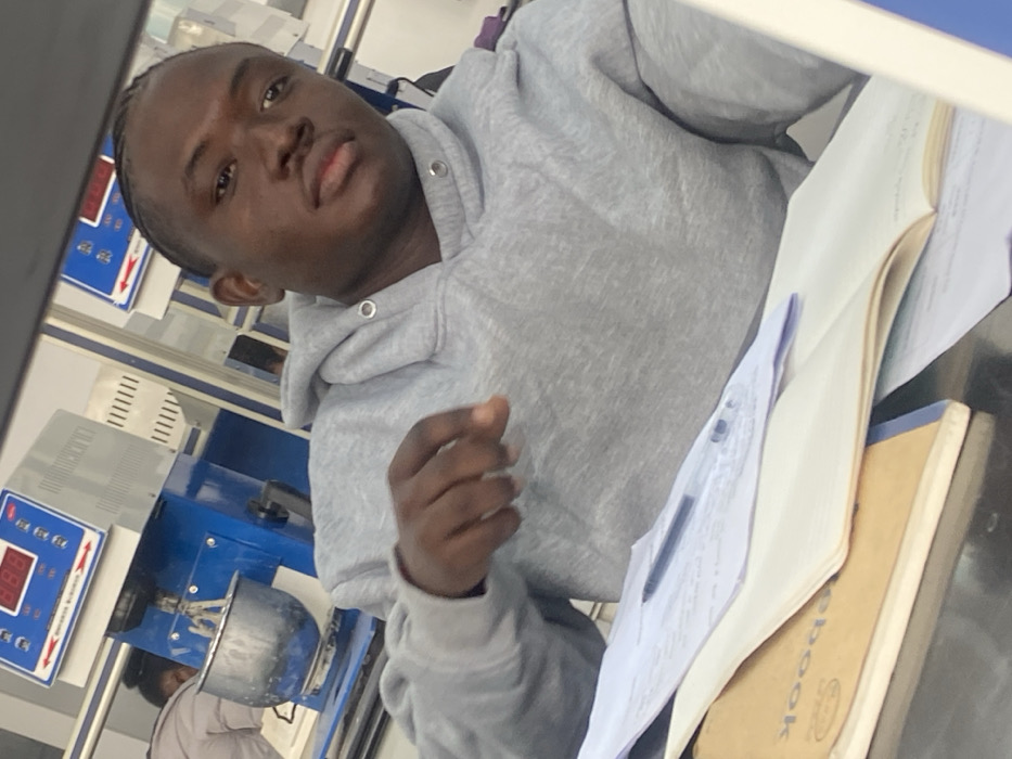A young man in a gray hoodie sits at a table with papers and a notebook. Behind him is lab equipment, including a machine with a digital display and a metal bowl. The man is facing the camera, and his hand is raised. Other lab equipment is visible in the background.