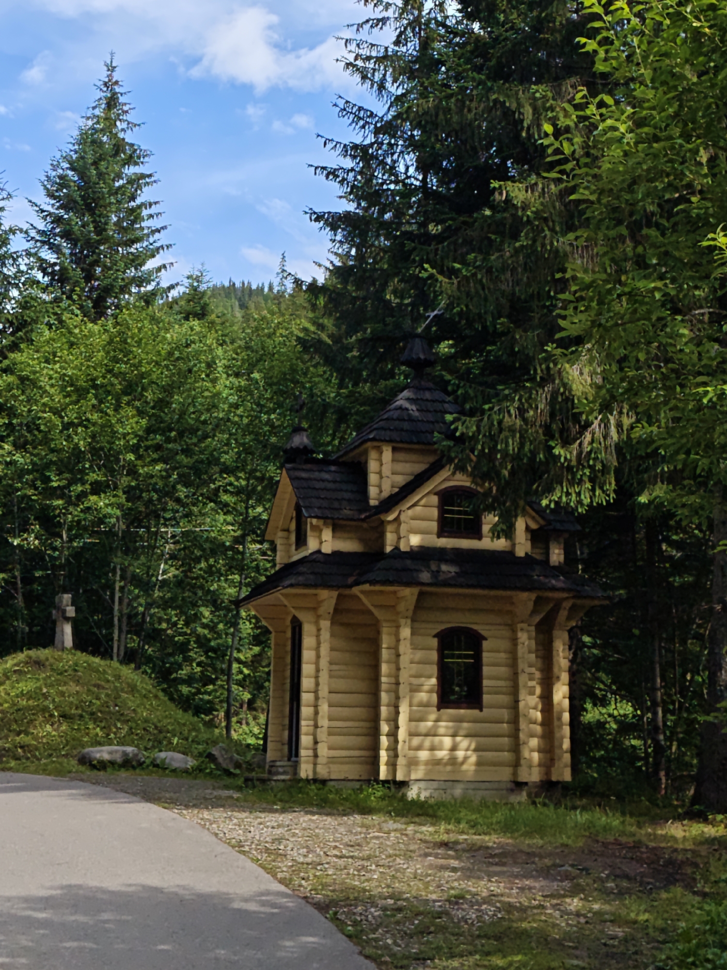 This image shows a small, wooden chapel situated amidst a dense forest in Tatariv, Ukraine. The chapel, constructed from light-colored logs, features a dark, multi-tiered roof with a central dome and smaller domes on the sides. It has arched windows with dark frames. To the left of the chapel, a grassy hill rises, topped with a stone cross. A paved path curves through the foreground, leading towards the chapel. The background is filled with various types of trees, predominantly conifers and deciduous trees, creating a lush green backdrop against a bright blue sky with scattered white clouds. The scene depicts a peaceful, natural setting, likely a religious or historical site.