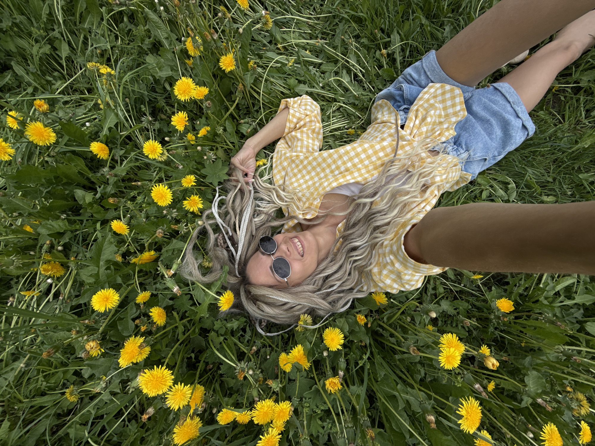 A young woman with long blonde wavy hair lies in a field of dandelions. She wears round sunglasses, a yellow and white checkered blouse, and denim shorts. Her arms are extended, holding the camera for a selfie. The field is green with numerous yellow dandelion flowers. The location is possibly in the city of Шебунино, Russia.