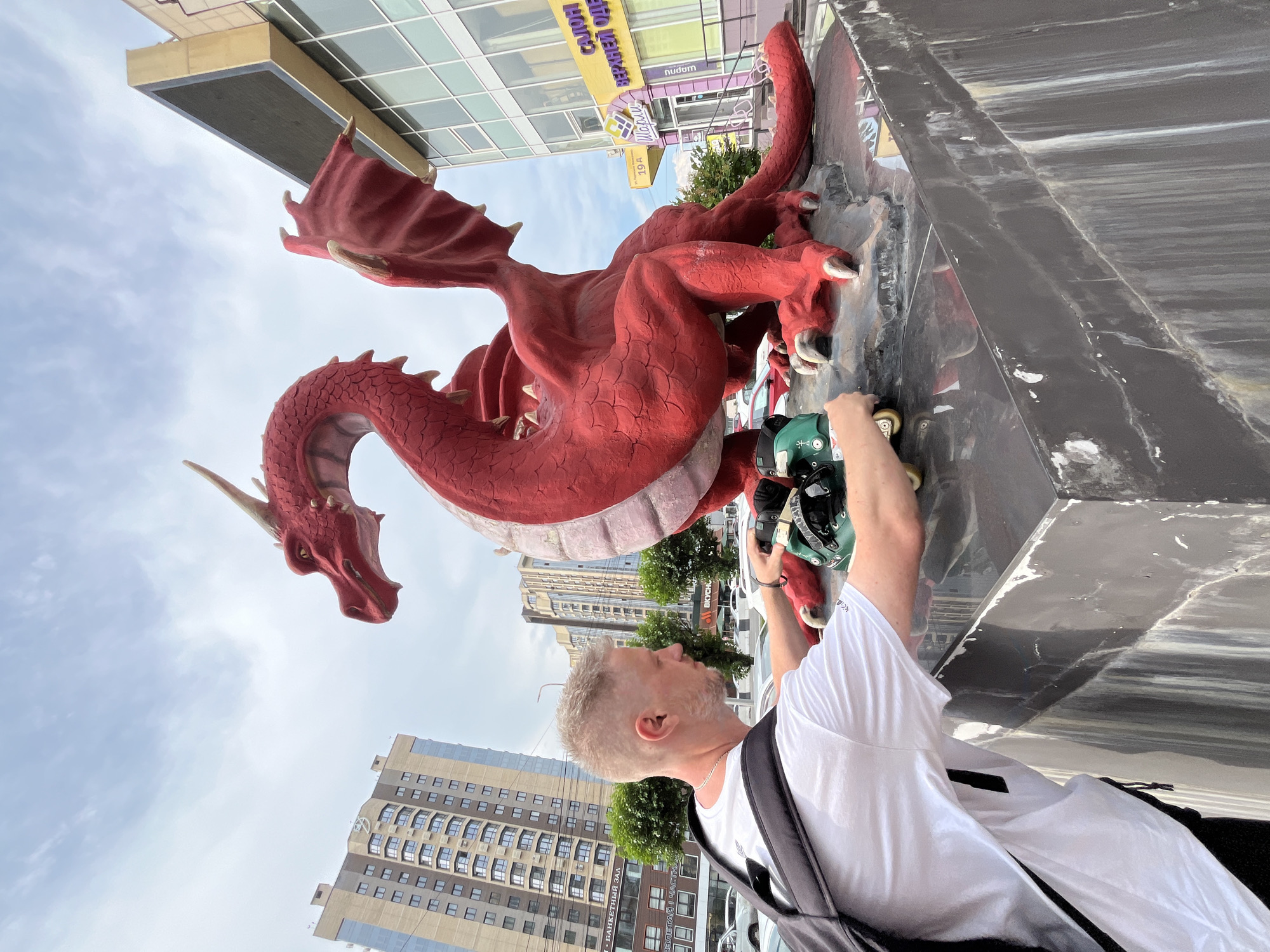 A blonde man wearing a white t-shirt and a black backpack is placing a pair of roller skates on a large, red dragon sculpture in the city of Krasnoyarsk, Russia. The sculpture is positioned in front of a modern building with a large digital billboard. The man is partially obscured by the sculpture as he leans forward to place the roller skates. The sky is overcast with clouds. In the background, there are several tall apartment buildings and trees. The sculpture is on a dark, reflective base.