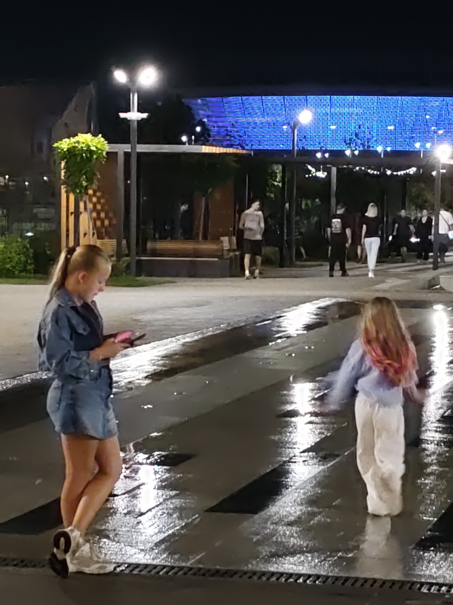 At night in Rostov-on-Don, Russia, two young girls are walking on a wet paved surface that reflects the ambient light from street lamps and a large blue LED screen overhead. The girl on the left, with blonde hair in a ponytail, is wearing a denim jacket and skirt and is looking at her phone. The girl on the right, with blonde hair streaked with pink, is wearing a light blue long-sleeved top and white pants and is facing away from the camera, with her arms outstretched as if dancing or running. In the background, several other people are visible, some walking and some standing, in an outdoor area that appears to be a public space or promenade. The overall atmosphere is lively and illuminated by artificial light.