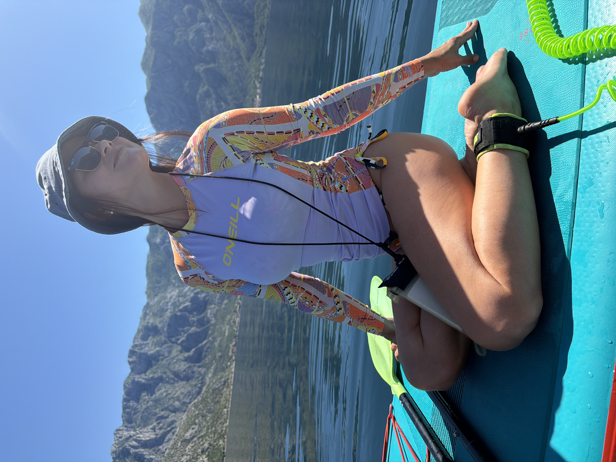 A woman wearing a hat and sunglasses is kneeling on a turquoise paddleboard on the water near the town of Stoliv, Montenegro. She is wearing a long-sleeved rash guard with orange and purple patterns, and her left foot is attached to the paddleboard with a black and green leash. A paddle is visible next to her, and mountains are visible in the background. The sky is clear and blue.