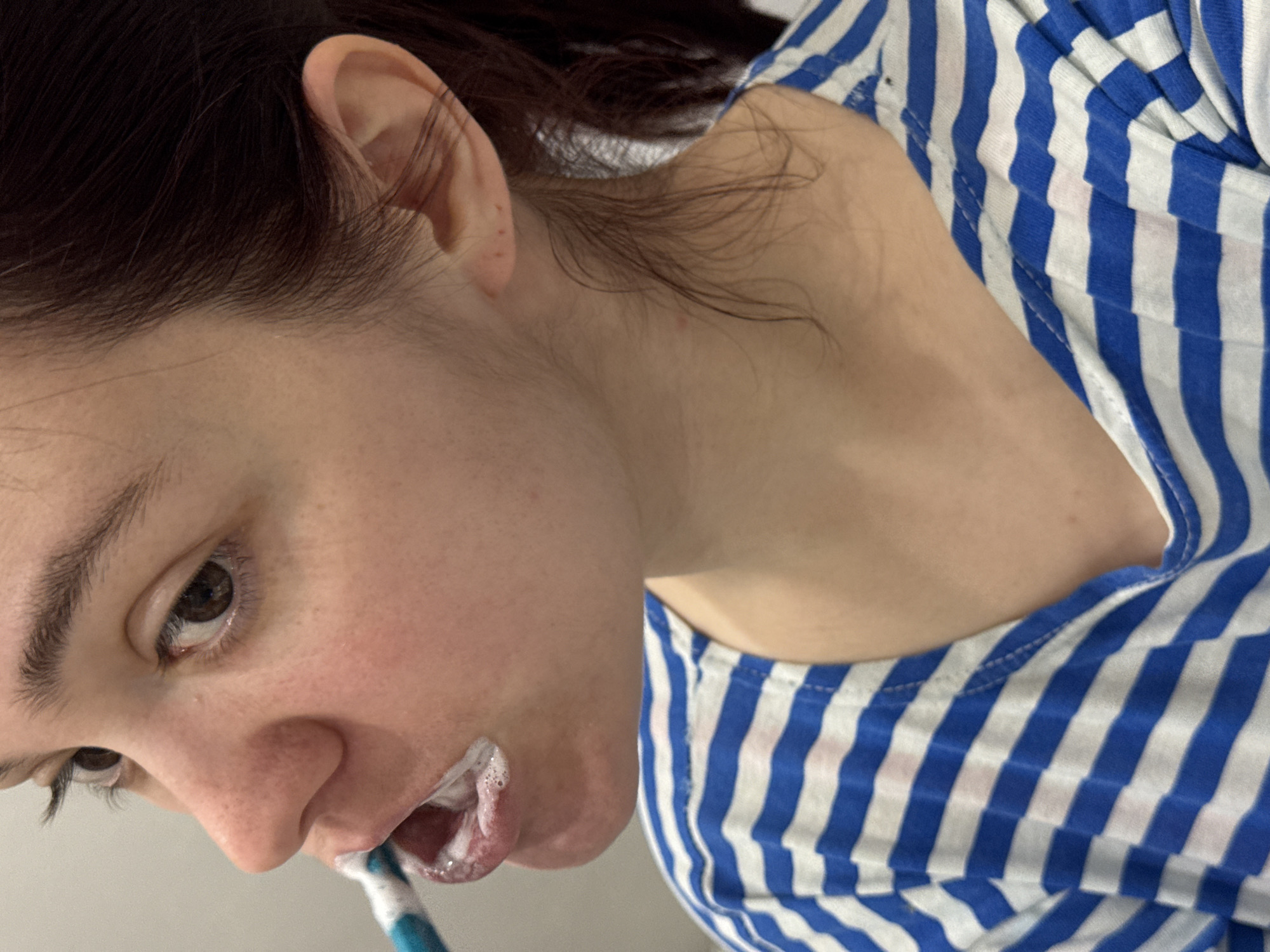 A close-up image captures a person, likely female, brushing their teeth in what appears to be an indoor setting, possibly a bathroom. The individual has dark brown hair, which is slightly pulled back, and fair skin, with a few small red spots visible on the ear and neck. They are wearing a blue and white striped top.
The person's head is tilted downwards and to the left, with their right eye visible and looking in the same direction, indicating focus on the task. A blue and white toothbrush is positioned in their mouth, surrounded by white toothpaste foam and suds. The background is a plain, light color, offering no specific environmental details. The lighting suggests an indoor setting, but the time of day is not discernible. No text is visible in the image, and no specific location cues are present.
