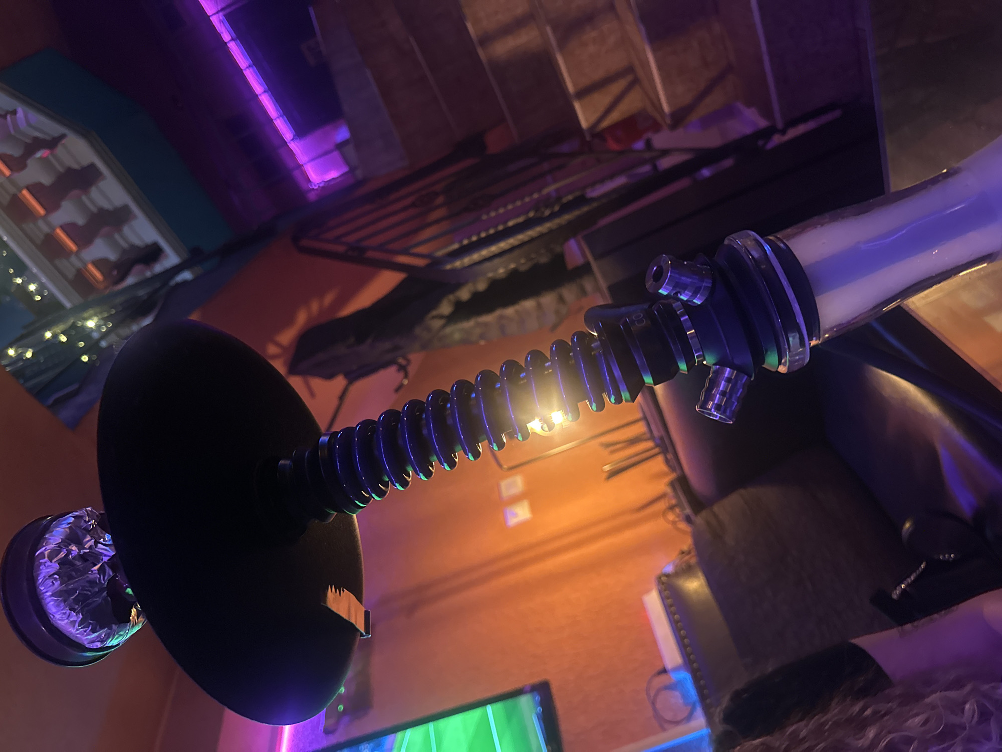 The image shows a hookah in a room with an orange wall, a black leather chair, and a television with a green screen. There is a metal shelf with lights above the hookah. The hookah has a black base, a silver-wrapped bowl, and a clear glass chamber filled with white smoke. The image does not contain enough details to determine the precise location in Varna, Bulgaria.