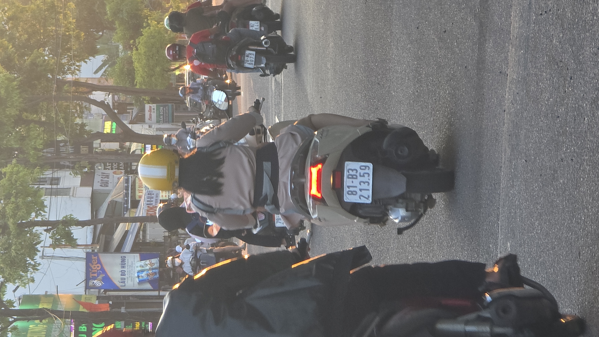 The image captures a street scene in an urban area of Vietnam, consistent with Nha Trang, during late afternoon or early evening, indicated by the warm, low-angle sunlight. A procession of motorbikes and their riders moves forward on an asphalt road.
The central subject is a woman riding a light brown or tan scooter, viewed from behind. She wears a yellow helmet with a white longitudinal stripe and a light brown long-sleeved top with a dark waist strap. Her dark hair extends down her back. The scooter's red taillight is illuminated, and its rear license plate, "81-B3 213.59", is clearly visible. To her immediate left, the rear of another dark-colored motorbike and its rider are partially visible.
Further into the background, a line of additional motorbikes and riders is present. Riders wear various helmets, including red and dark colors, and some are observed wearing face masks. Visible license plates on other scooters include "78-M1 9073" and "79-NZ 8.44". The plate "79-NZ" is consistent with Khanh Hoa province, which includes Nha Trang, while "81-B3" corresponds to Gia Lai province and "78-M1" to Phu Yen province.
The environment consists of multi-story buildings lining the street, partially obscured by green-leafed trees. Prominent signage in Vietnamese includes a large blue "Tiger" beer banner advertising "LẨU BÒ HÙNG" (Beef Hotpot Hung) and a sign reading "GIẶT SẤY" (Laundry). Overhead power lines are visible across the top of the frame.