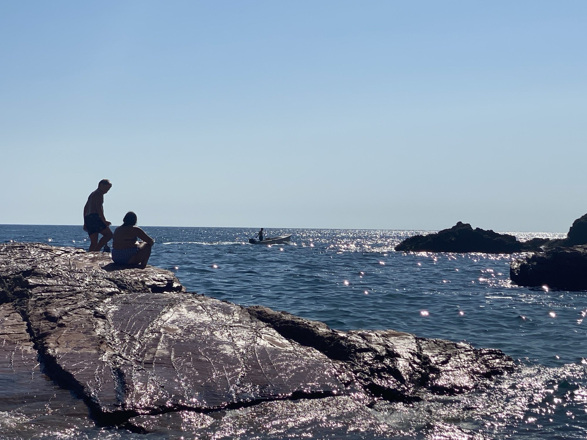 In the city of Sutomore, Montenegro, this photograph captures a serene coastal scene under a clear, bright sky. The foreground is dominated by large, dark, textured rocks, glistening with the reflection of sunlight on their wet surfaces. Two individuals are silhouetted on a rock ledge in the mid-left of the frame, one standing and the other seated. The standing figure, appearing to be male, is wearing shorts. The seated figure, appearing to be female, is wearing swimwear. Beyond the rocks and people, a vast expanse of deep blue water stretches towards the horizon, dotted with sparkling reflections of sunlight. In the mid-distance, a small motorboat with a single occupant is moving across the water, leaving a subtle wake. Further out to sea, several dark rocky islets rise from the water. The overall impression is one of a tranquil summer day by the Adriatic Sea.