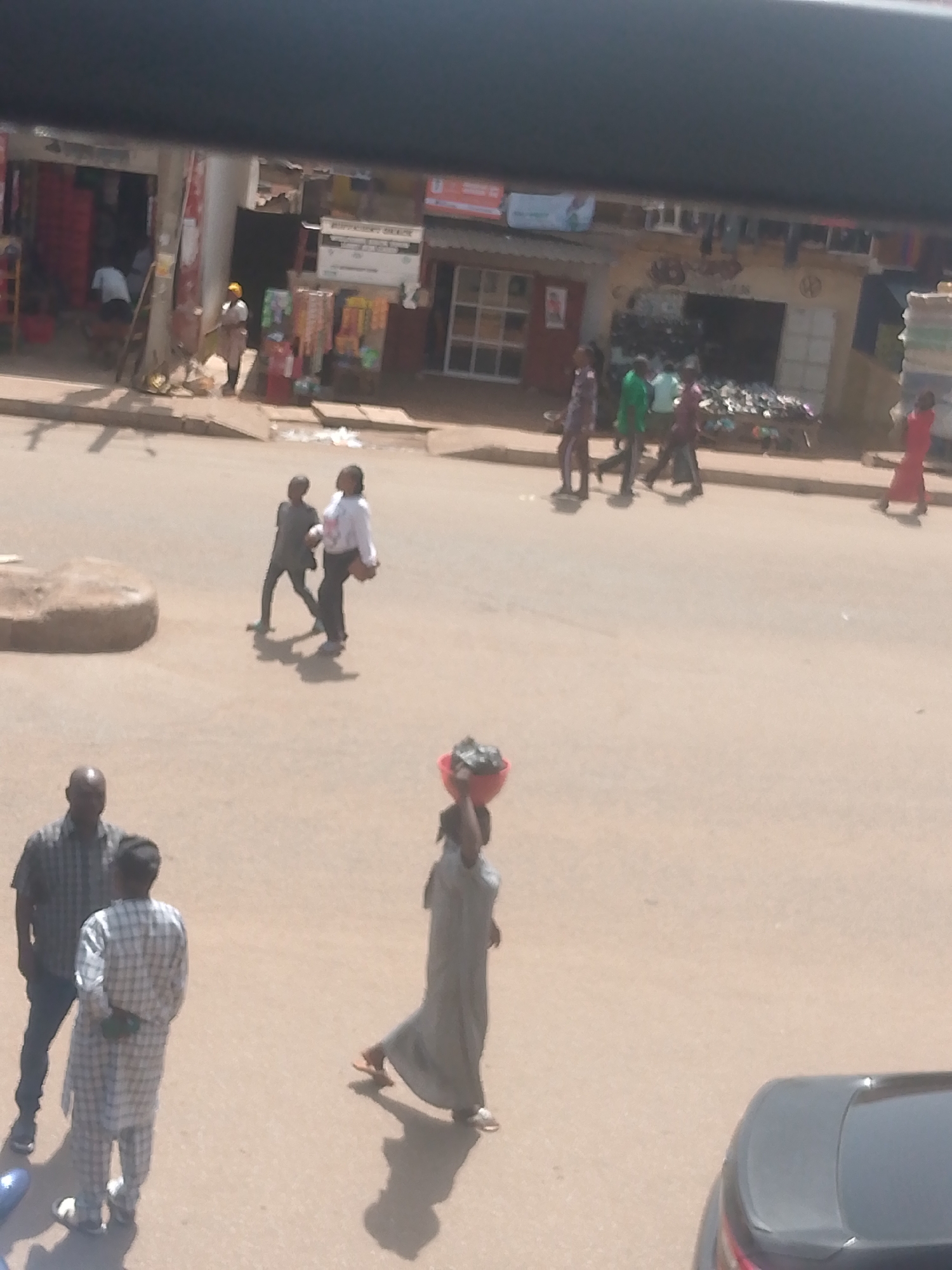 The image depicts a street scene in Bukuru, Nigeria, with several people walking and interacting. In the foreground, two men stand on the left, one wearing a checkered shirt and pants, and the other a dark checkered shirt with blue jeans. To the right of them, a woman in a long grey garment walks, carrying a red bowl on her head. In the mid-ground, two figures are walking away from the viewer, one in a dark outfit and the other in a white shirt and dark pants, possibly carrying a bag. Further back, a group of people are walking on the street, including individuals in green and dark red clothing. The background shows a row of buildings with shopfronts, displaying various items, including clothing and other goods. There appears to be some construction or debris in front of the shops. The scene suggests a busy marketplace or commercial area. The lighting indicates it is daytime, possibly late morning or afternoon, with shadows cast on the ground. No specific event is apparent, other than general daily activity. There is no visible text in the image that is clear enough to be read.