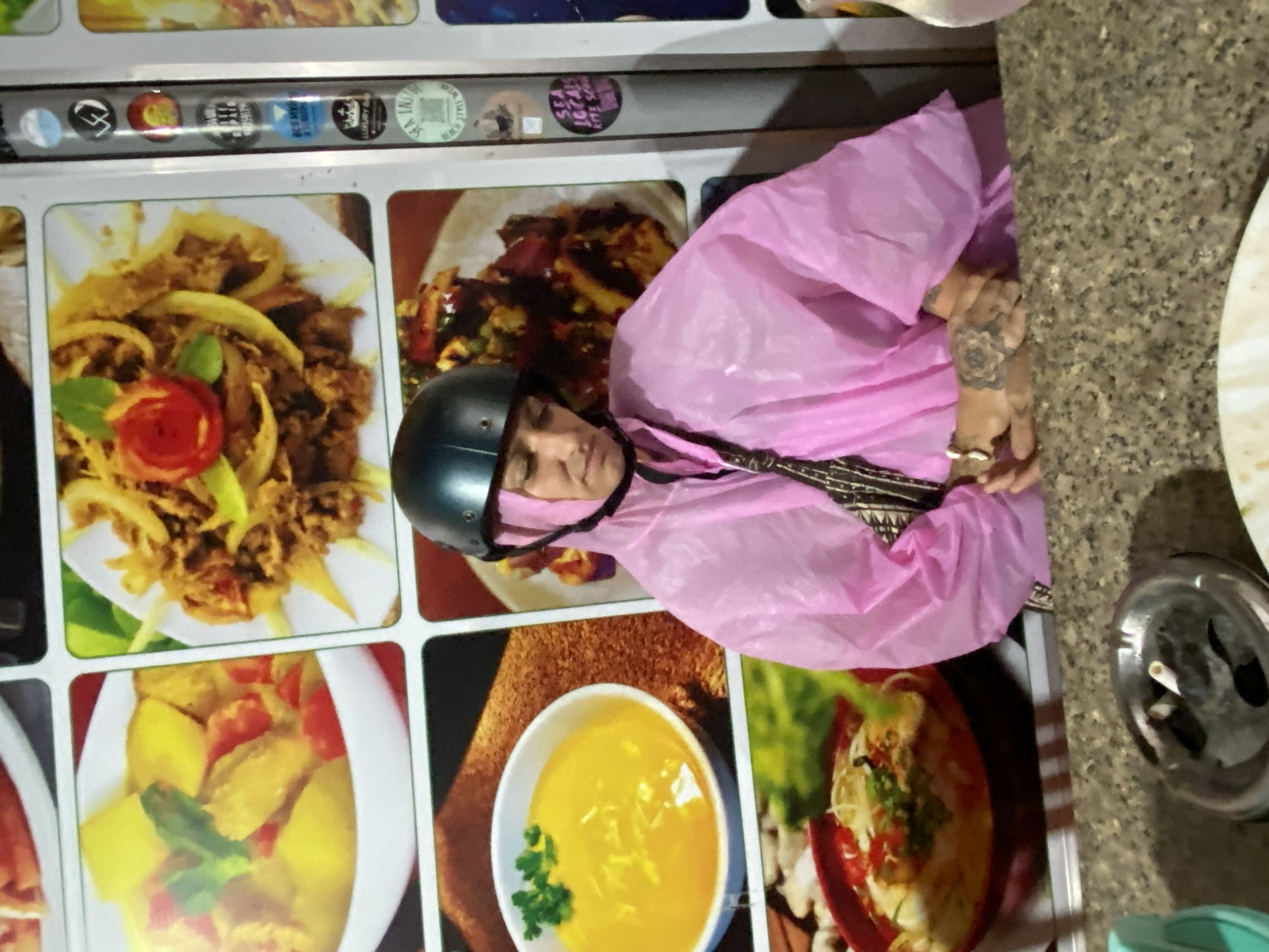 The image captures a scene in what appears to be a casual eatery or food stall, likely in Phu Thuy Ward, Vietnam. The central subject is a person, seen from a slightly elevated angle, wearing a black motorcycle helmet and a bright pink plastic rain poncho, indicating rain or recent wet weather. The person's hands are visible beneath the poncho, adorned with intricate tattoos, including a prominent mandala-like design on the back of the left hand, and several rings. Their expression is neutral as they look downwards or straight ahead.
To the left, a large display panel functions as a menu, featuring multiple square images of food dishes. Visible dishes include a stir-fried meal with onions and meat, a yellow curry-like dish containing potatoes and tomatoes, and a bowl of yellow soup garnished with herbs. Above this food display, a narrow shelf holds several circular items, possibly stickers or bottle caps, bearing text such as "SPA JESSICA", "KIT SON", "BE MY", "LUXURY", "SAI BEACH", and "JAI TAI", which seem to be local advertisements or brand names.
To the right of the person, a speckled, possibly granite, counter or table surface is visible, on which a metallic ashtray containing several cigarette butts rests. The indoor lighting suggests the time of day could be evening. The attire of the person, combined with the food display and ashtray, points to a typical informal dining experience common in Vietnam.