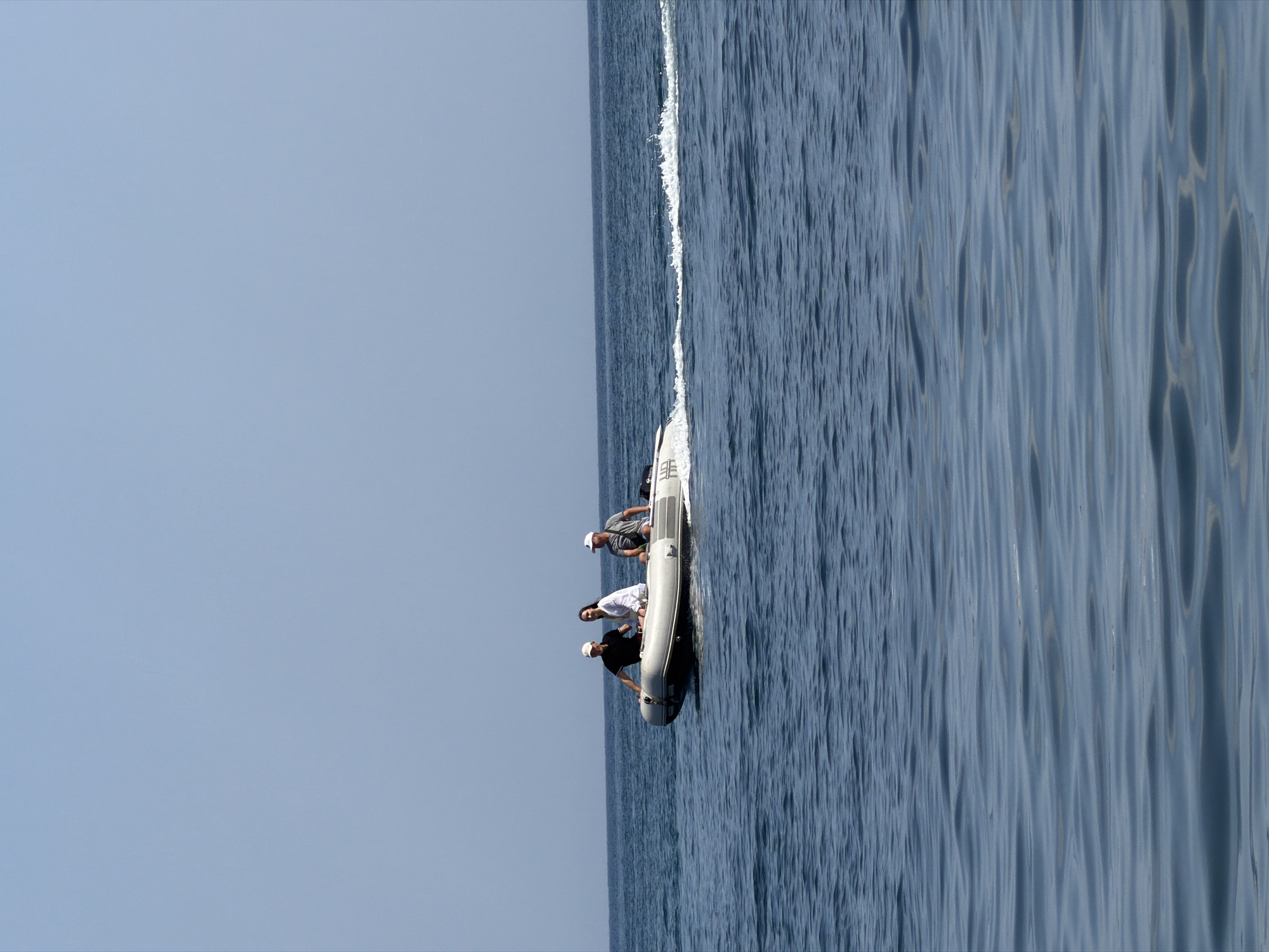 An inflatable boat with three people aboard travels across a body of water under a clear sky. The boat is white with a dark gray stripe along its side. A man in a black shirt and white cap is at the helm, steering the boat. Next to him, a person in a white shirt and a long-haired individual in a white shirt and dark pants are seated. On the right side of the boat, a man in a gray shirt and white cap is seated. The boat is creating a wake behind it. The water is calm with gentle ripples. The horizon line where the water meets the sky is visible in the distance.