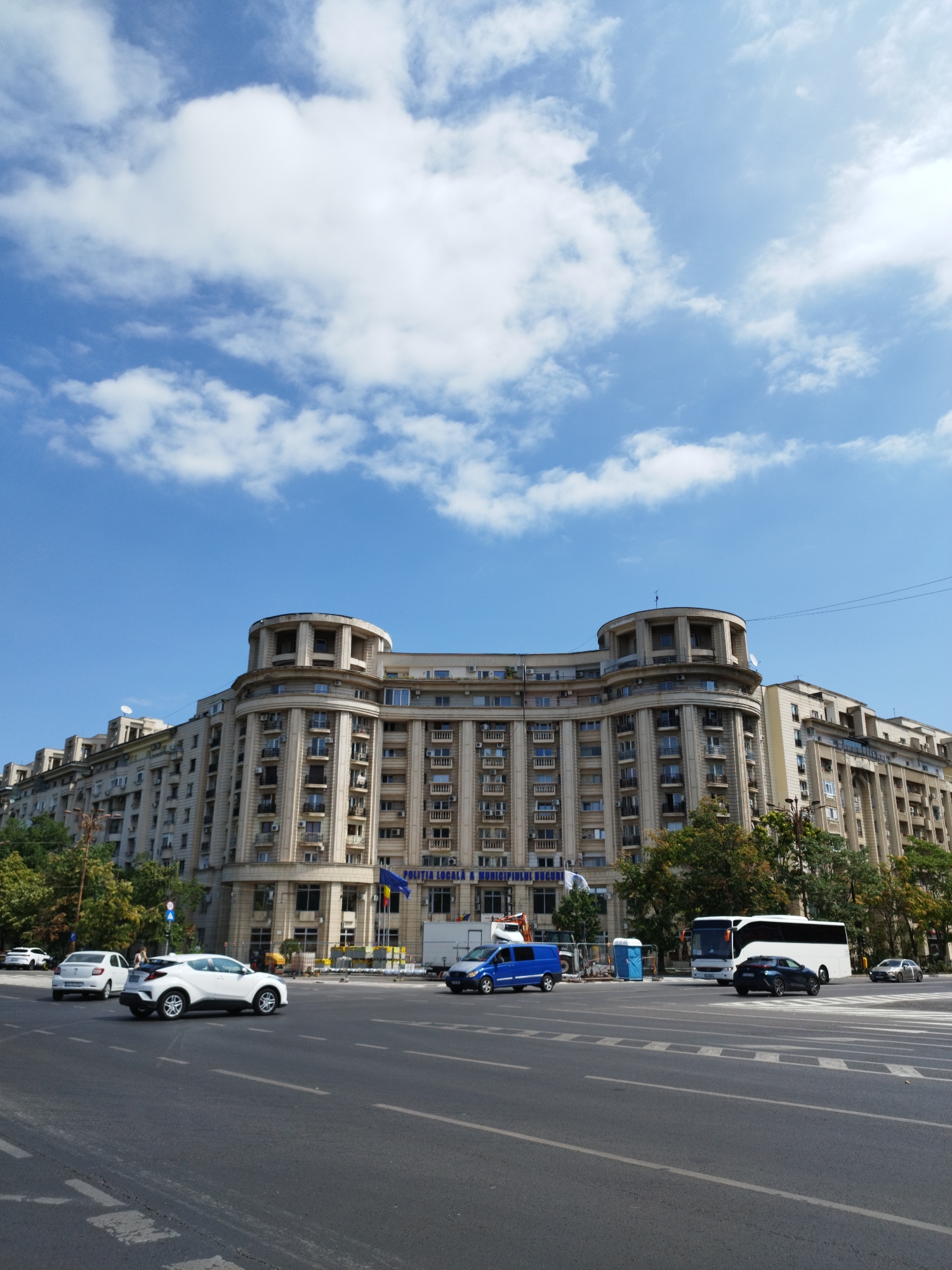This image depicts a street scene in Bucharest, Romania, featuring a large, multi-story building with a curved facade, characteristic of early 20th-century architecture. The building is light-colored, with numerous windows and balconies, and is adorned with classical architectural elements like columns and decorative moldings. A blue banner with Romanian text is visible on the building, likely indicating an official institution.
The foreground shows a multi-lane road with several vehicles, including cars and a white bus. The road surface is asphalt, marked with white lines indicating lanes and a pedestrian crossing. The sky above is blue with scattered white clouds. Trees line the street, providing a touch of greenery. Construction barriers and portable toilets are present on the side of the road, suggesting ongoing work in the vicinity. The overall impression is of a bustling urban environment under a clear sky.