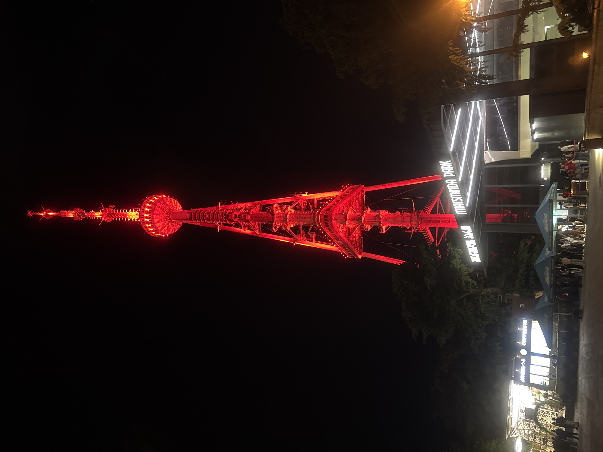 The image shows the Mtatsminda Park TV tower in Tbilisi, Georgia, illuminated in red at night. The tower is a tall, slender structure with a distinctive red glowing crown and upper section. Below the tower, there is an entrance area with a sign that reads "MTATSMINDA PARK". People are visible gathered in front of the entrance, some queuing and others standing around. There are also some trees and a building with lit-up windows, possibly a cafe or shop, to the left of the entrance. The overall scene depicts a popular nighttime tourist attraction.