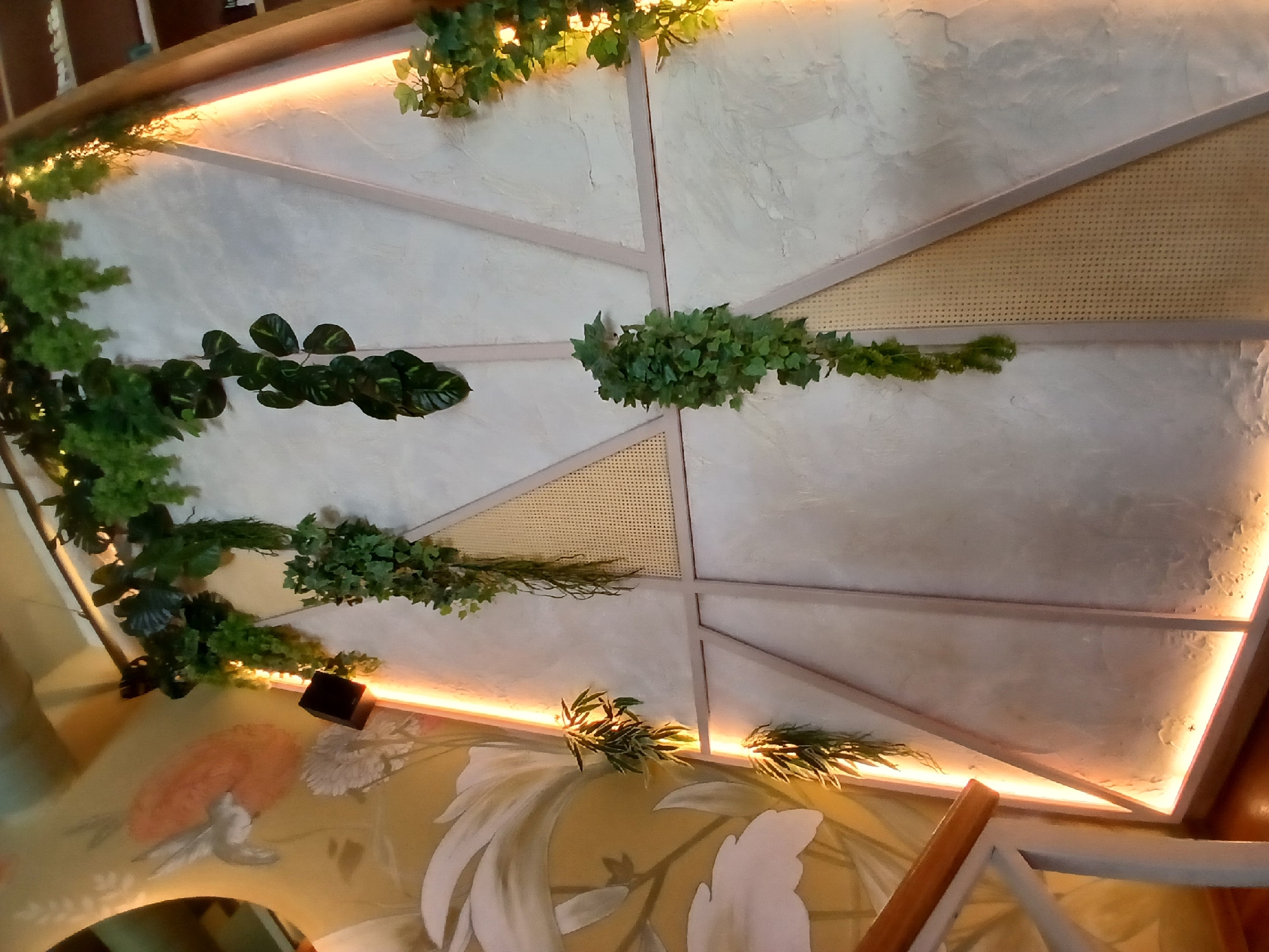 The image shows a wall decorated with hanging plants and geometric panels. A staircase railing is visible in the lower left corner. The wall features a mural on the left side depicting a bird and floral elements. The wall itself is textured and has several triangular panels, some with a woven cane material. Strip lighting is integrated along the edges of some of the panels, casting a warm glow. The style of the decor suggests it could be an interior space like a cafe, restaurant, or a home. There is no information in the image to pinpoint its location in Yekaterinburg, Russia.