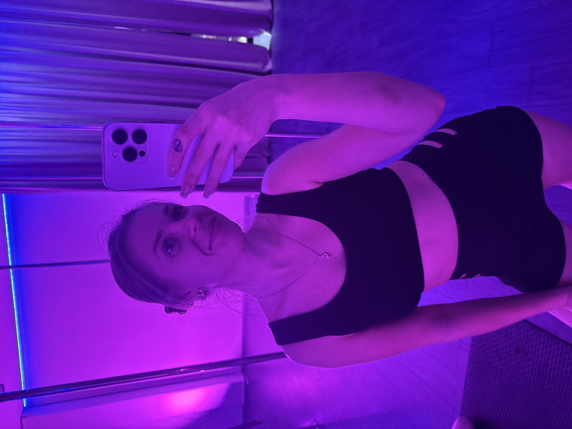 A woman is indoors, taking a selfie with her smartphone. The phone is held up in her left hand, revealing multiple camera lenses on the back. She has a small tattoo on her left index finger. She is wearing a black athletic top and matching shorts with cut-out designs. The lighting is predominantly purple, suggesting a tanning bed environment. The background shows horizontal metal bars. The location is a tanning salon. It is not possible to know that the tanning salon is in the city of Batumi, Georgia, given the lack of location indicators in the image.