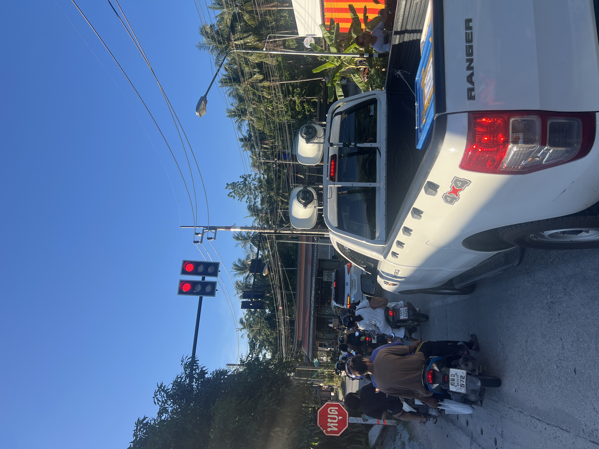 In the town of Koh Phangan, Thailand, a traffic jam unfolds under a clear blue sky. A white Ford Ranger pickup truck, adorned with "4x4" and "RANGER" badging, is prominently featured in the foreground, its red brake lights illuminated. The truck bed contains a blue plastic tray with items inside. Ahead of the truck, a line of motorcycles and their riders are stopped. The riders, mostly wearing helmets and casual attire, are patiently waiting. One motorcycle displays the license plate "103 4429". Further ahead, a white SUV with license plate "790 4602" is also stopped. Traffic lights above the intersection show red lights for all directions. To the left, a red octagonal "STOP" sign with Thai lettering is visible. The background is filled with lush green trees, including palm trees, and overhead electrical wires are strung across poles. The scene appears to be a typical roadside setting in a tropical location.