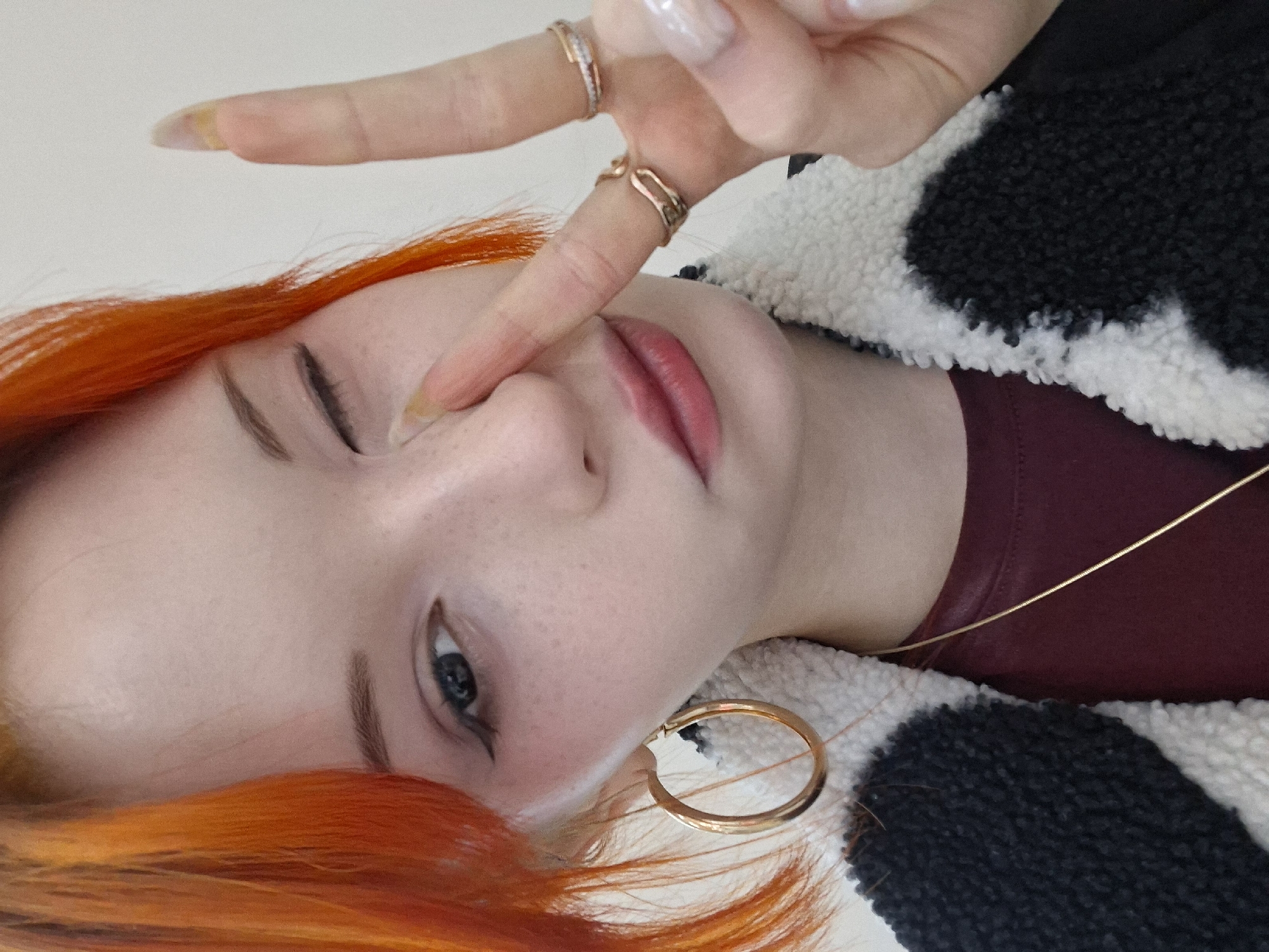 A close-up, portrait-oriented image shows a young woman with orange hair making a peace sign. The woman has light skin with freckles, one visible blue eye, and pink lips. She is wearing a large gold hoop earring in her left ear. On her fingers, she is wearing multiple gold rings. She is wearing a black and white, sherpa-style jacket over a maroon shirt. A thin gold necklace is visible around her neck. The background appears to be a solid white color. The image does not provide any information about a location in the city of Chaikovsky, Russia.