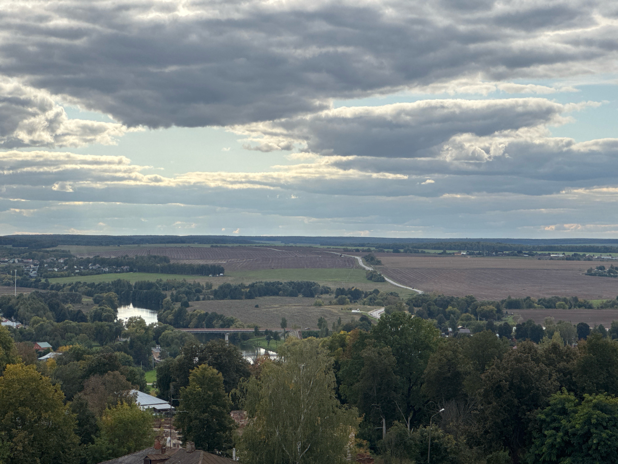 This aerial, wide-angle photograph captures a panoramic view of the city of Zaraysk, Russia, likely taken from a high vantage point such as a church bell tower or a hill. The sky is dominated by a dramatic arrangement of dense, dark grey clouds interspersed with brighter, lighter grey and white cloud formations, hinting at an overcast or stormy weather condition. Sunbeams break through in places, illuminating sections of the landscape.
The foreground features a cluster of red-tiled roofs belonging to buildings, partially obscured by lush green and autumnal trees with some leaves displaying yellow and brown hues. A streetlamp is visible amongst the trees.
The midground reveals the city's layout with more houses and buildings nestled among dense trees. A winding river, reflecting the sky, flows through the scene, crossed by a red bridge. The riverbanks are lined with trees and some agricultural land.
The background presents a vast expanse of cultivated fields and meadows, stretching towards the horizon. A road is visible snaking through the agricultural landscape. In the far distance, a faint treeline marks the edge of the terrain under the cloudy sky. The overall impression is of a rural or semi-rural Russian landscape under a dynamic sky.