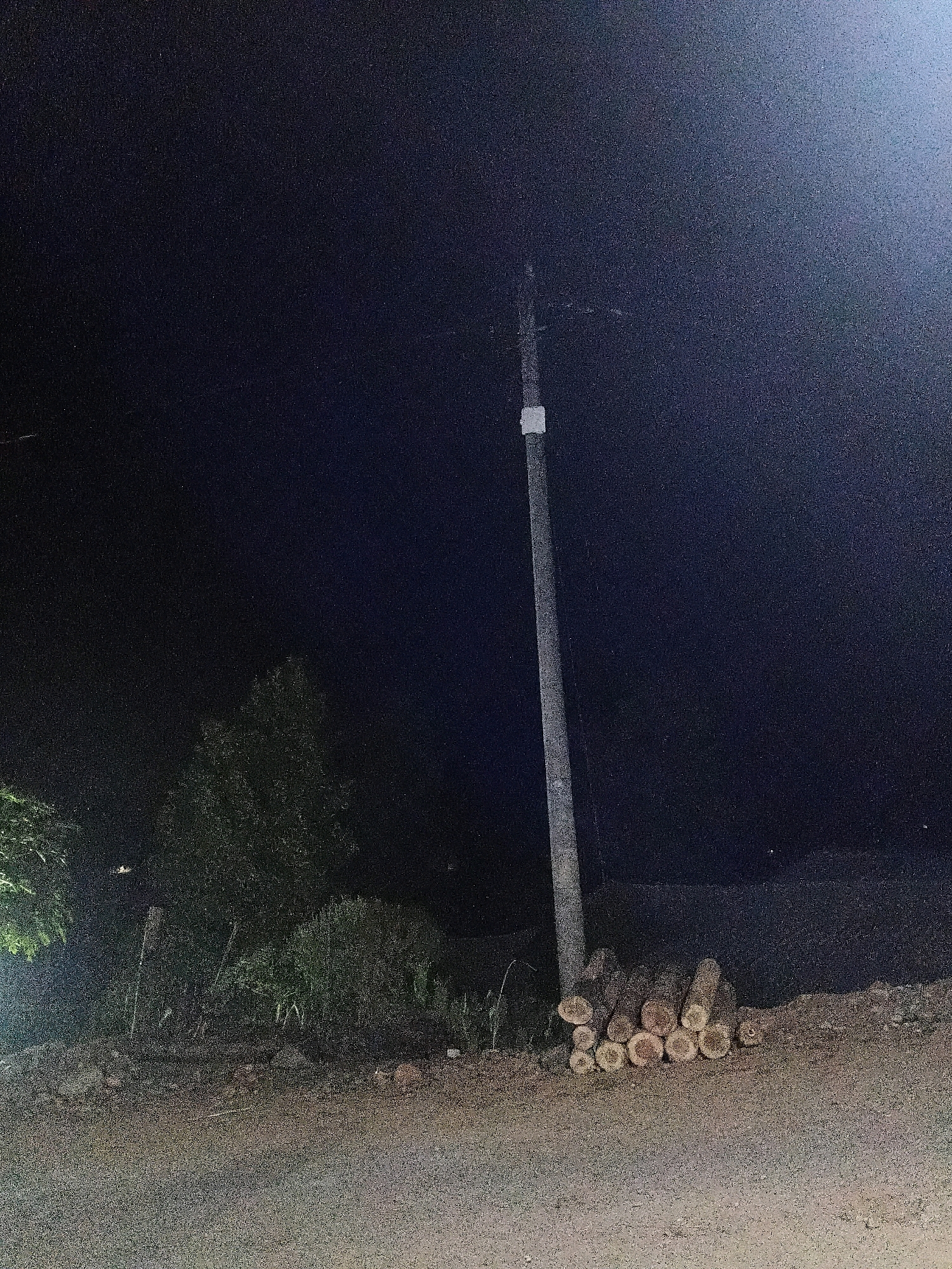 A nighttime photograph captures a rural scene in Sinduaji, Indonesia, featuring a solitary utility pole standing tall against a dark, star-speckled sky. The pole, a light gray cylinder, is positioned slightly off-center to the right of the frame. At its base, a neatly stacked pile of cut logs rests on the rough, earthy ground. To the left of the pole, dense green foliage, likely trees and bushes, creates a dark, natural backdrop. The foreground is occupied by a dirt path, its surface uneven and textured. The overall impression is one of quiet isolation in a rural Indonesian setting.