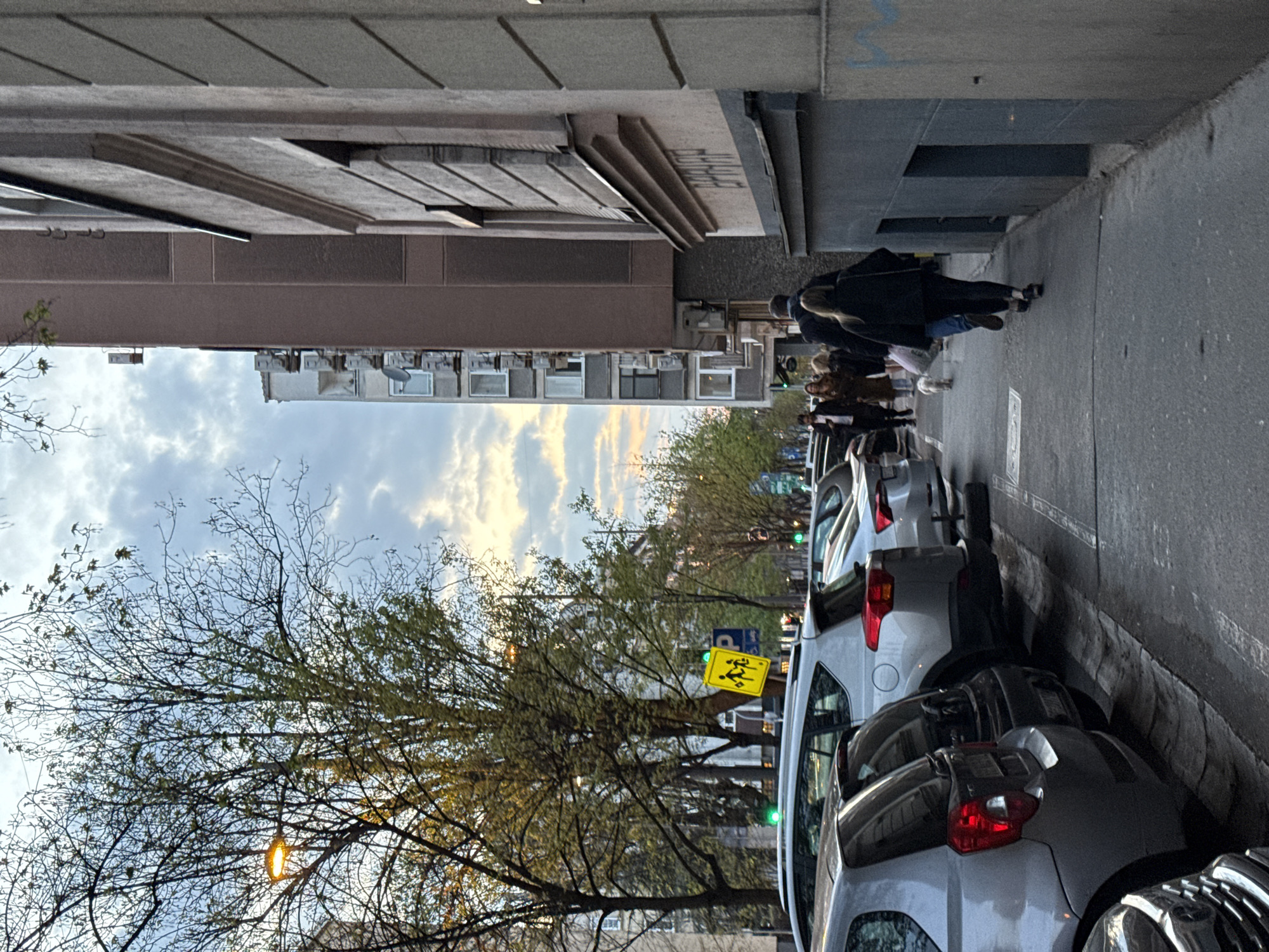 The image shows a street scene in Belgrade, Serbia. The viewpoint is looking down the street, between buildings on either side. There are several parked cars on the right side of the street. Pedestrians, including a woman with a dog, are walking on the sidewalk. A street sign with the image of children crossing is visible amongst bare trees. The sky is cloudy, with a hint of sunset.