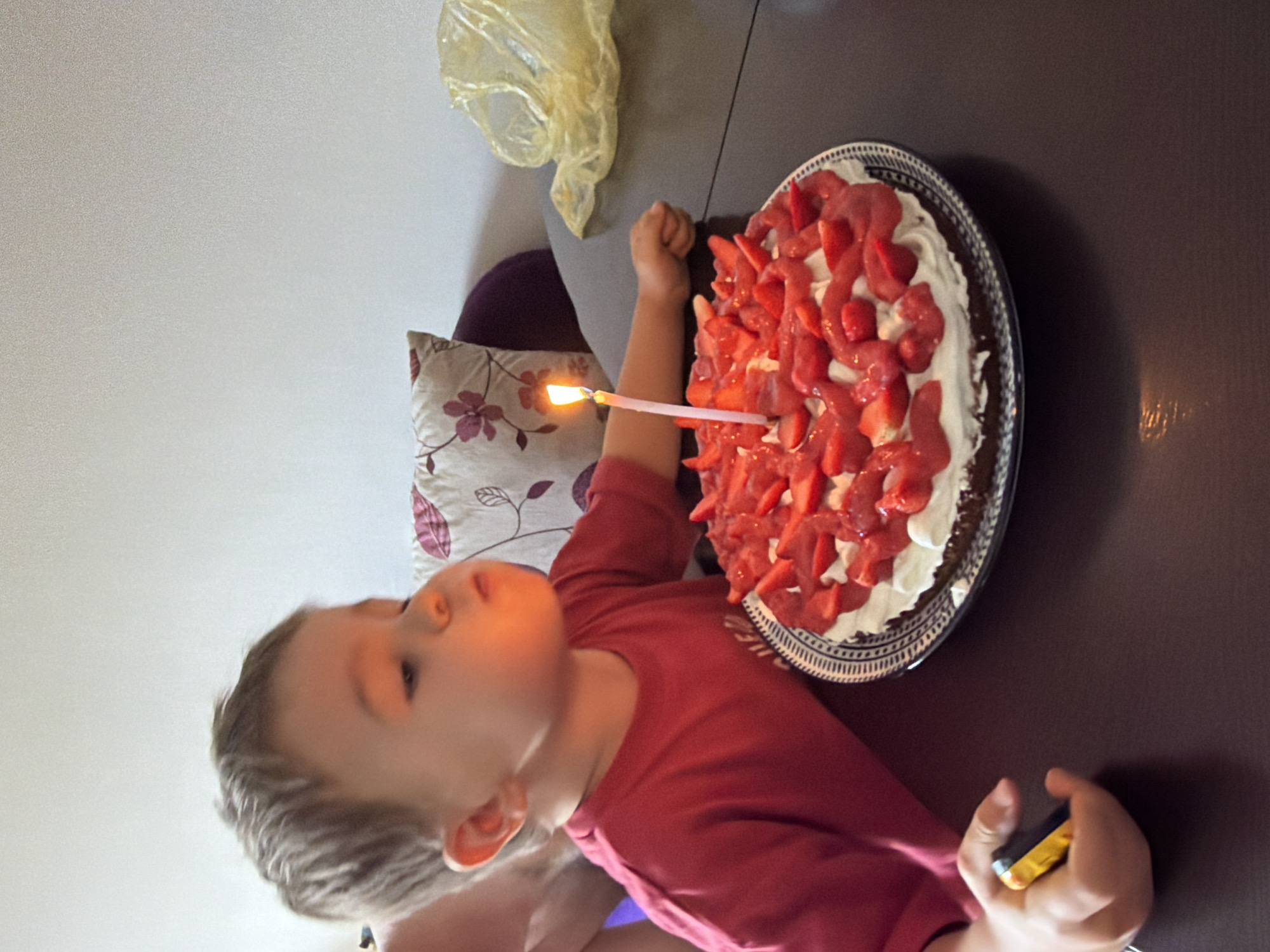 A young child in a red shirt is facing a strawberry-topped cake with a lit candle. The cake rests on a plate on a dark surface. The child is holding a small, dark object. A decorative pillow with floral patterns is visible behind the child. The setting is indoors, with a cream-colored wall in the background. There is no information available to conclude that the image was taken in Budva, Montenegro.