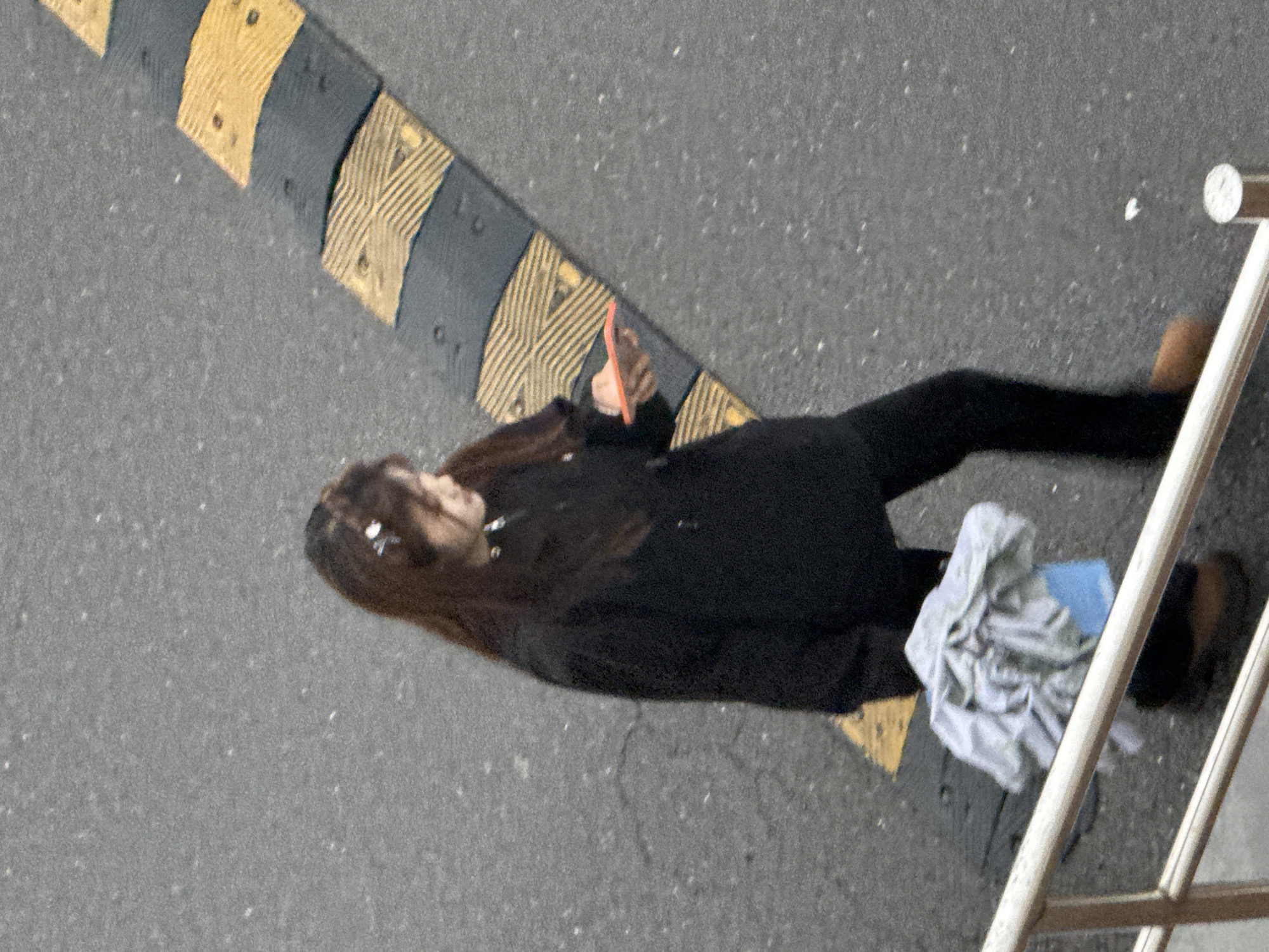The image, taken from an elevated perspective, shows an unidentified person standing on asphalt pavement in an urban environment. The person is wearing dark clothing and holding a small, possibly pink, rectangular object in their hand. To the left of the person, there is a section of a tactile paving surface with alternating yellow and dark grey stripes. To the right of the person is a set of white metal railings, with a crumpled white cloth and a blue object on the ground beside them. The background appears to be a gray asphalt surface. Location information suggests this scene is in Zhongtun, China.