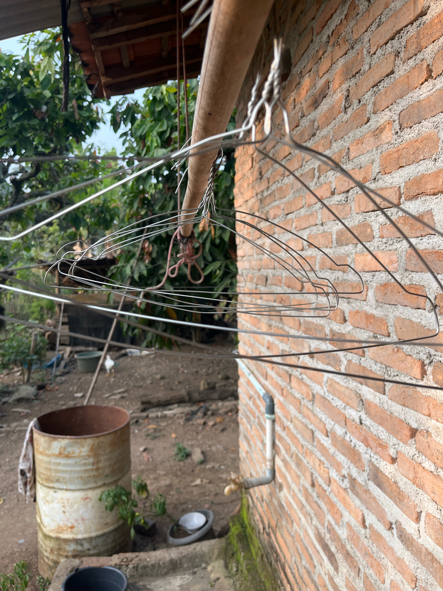 The image depicts an outdoor scene in Kebumen, Indonesia, featuring a clothesline strung with several wire hangers. The clothesline is suspended from a wooden beam that runs horizontally across the frame. To the right of the clothesline is a brick wall, constructed from orange and red bricks with gray mortar. A white pipe with a faucet is visible on the lower right portion of the brick wall. In the background, green foliage from trees and plants is present. On the left side of the image, a rusty metal drum stands upright, with a piece of fabric draped over its rim. The ground appears to be dirt and gravel, with some scattered debris. A small white chicken is visible in the middle ground. The overall impression is of a humble, rural or semi-rural setting.